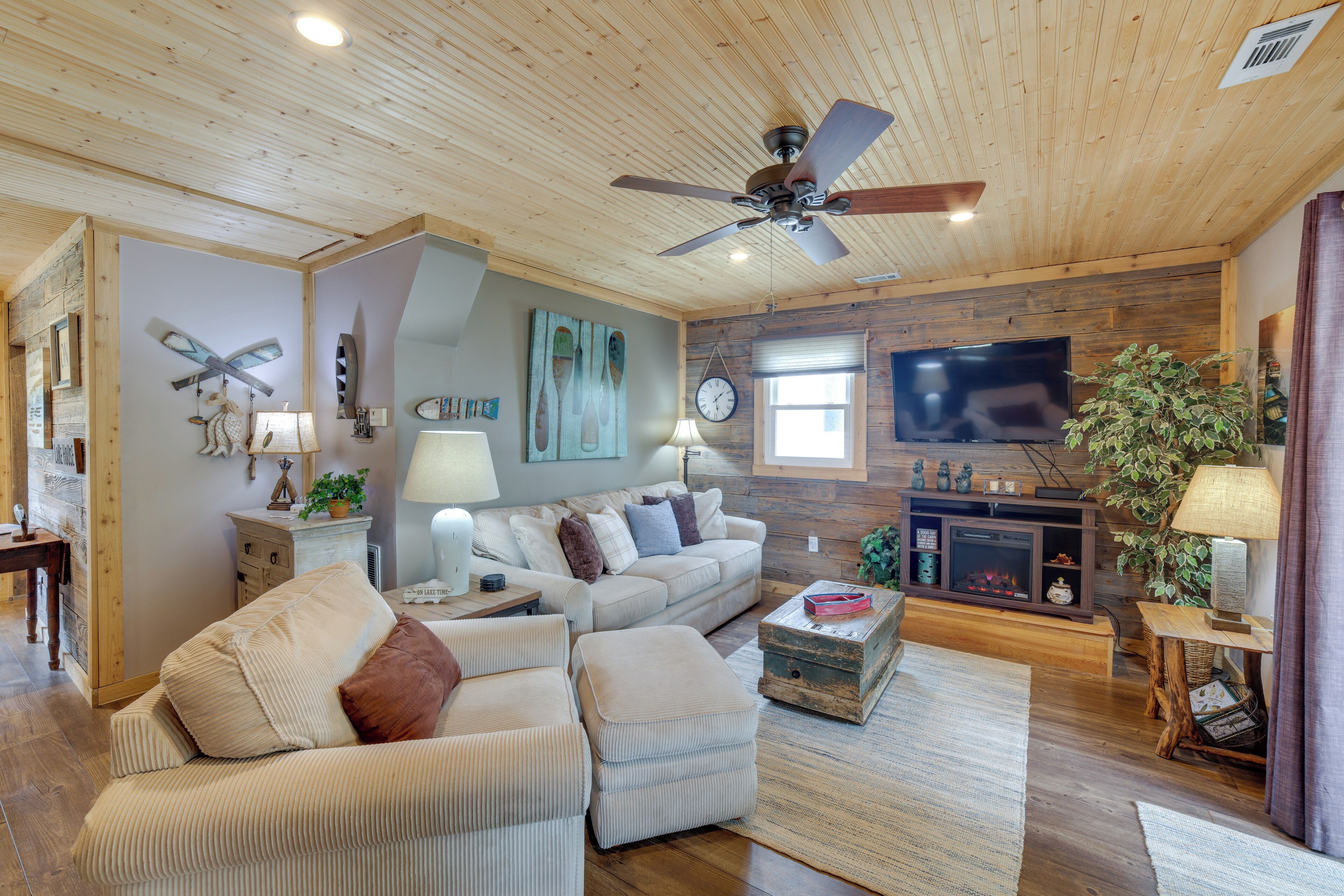A cozy living room with a wooden ceiling, a ceiling fan, a flat-screen TV, a fireplace, and a comfortable seating area.