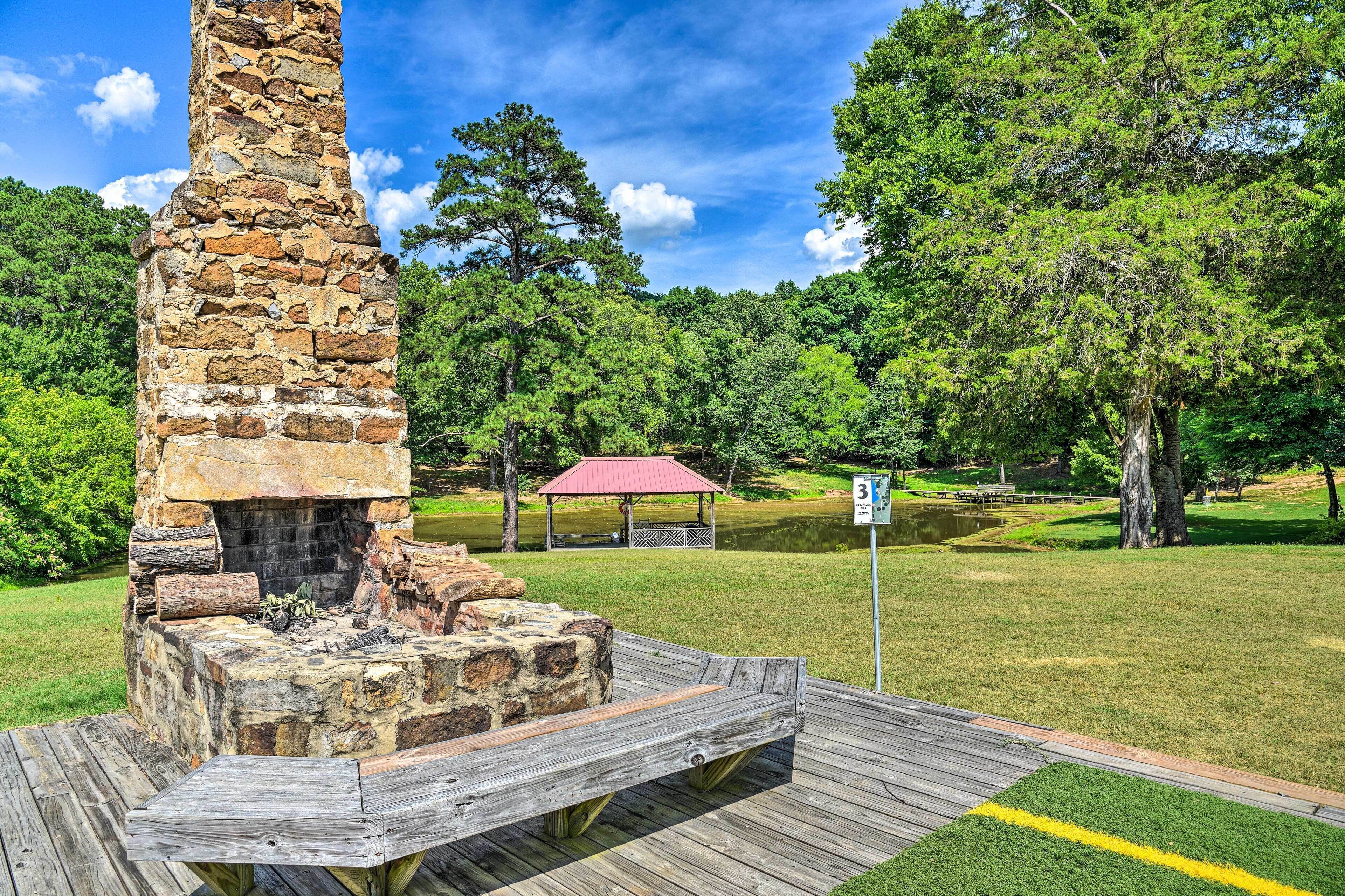 Una chimenea de piedra con una terraza de madera y mesa de picnic, rodeada por un parque con un pabellón y un estanque.