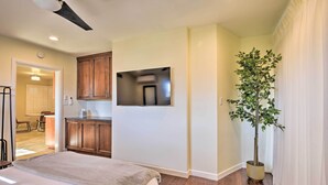 A bedroom with a bed, a ceiling fan, a TV mounted on the wall, wooden cabinets, a potted plant, and a view into the adjacent kitchen.