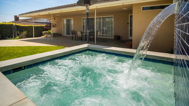 A swimming pool with a water feature, a covered patio with chairs, and a building with a tiled roof.