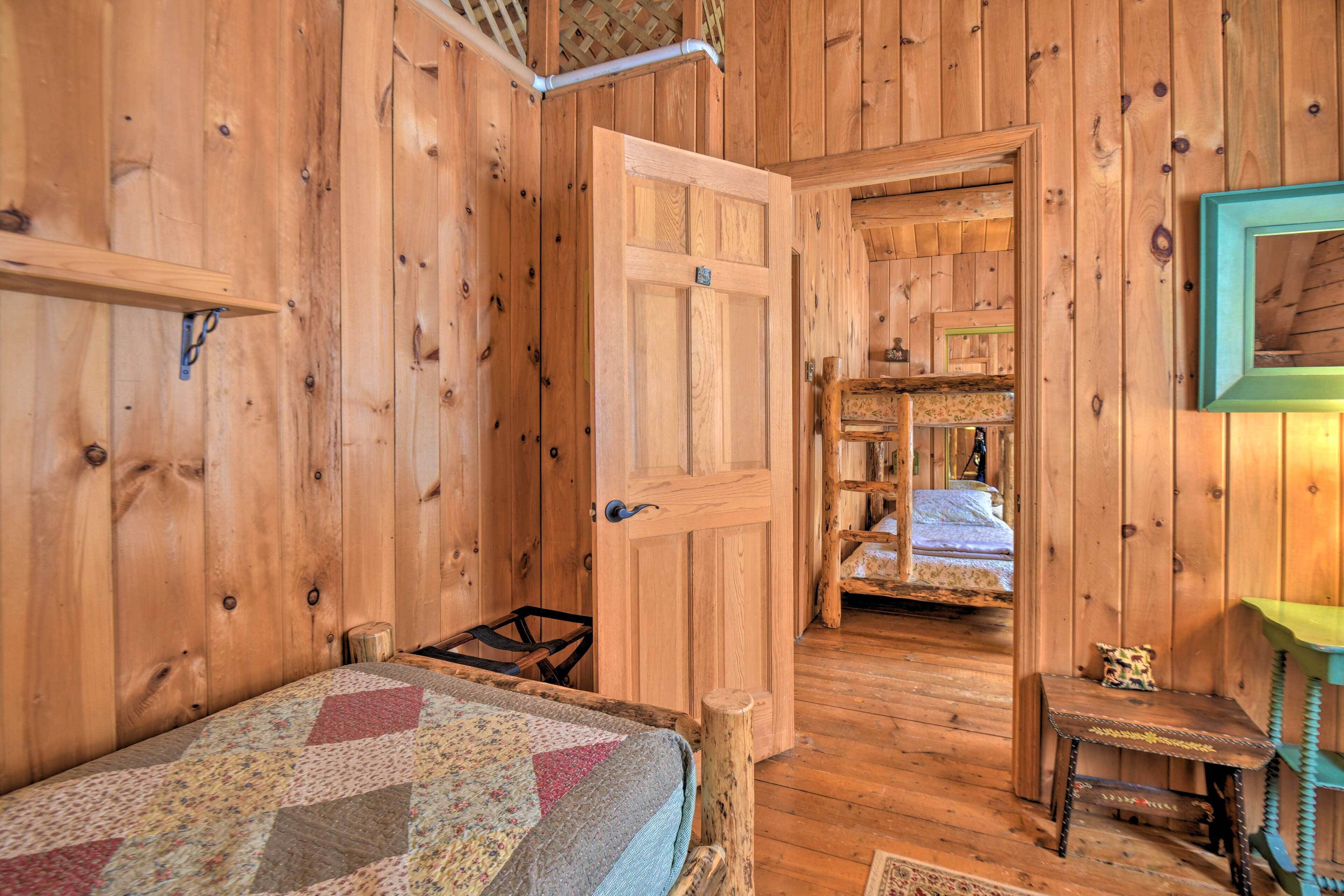 A wooden cabin room with a bed, a door leading to another room, and a small table with a lamp.