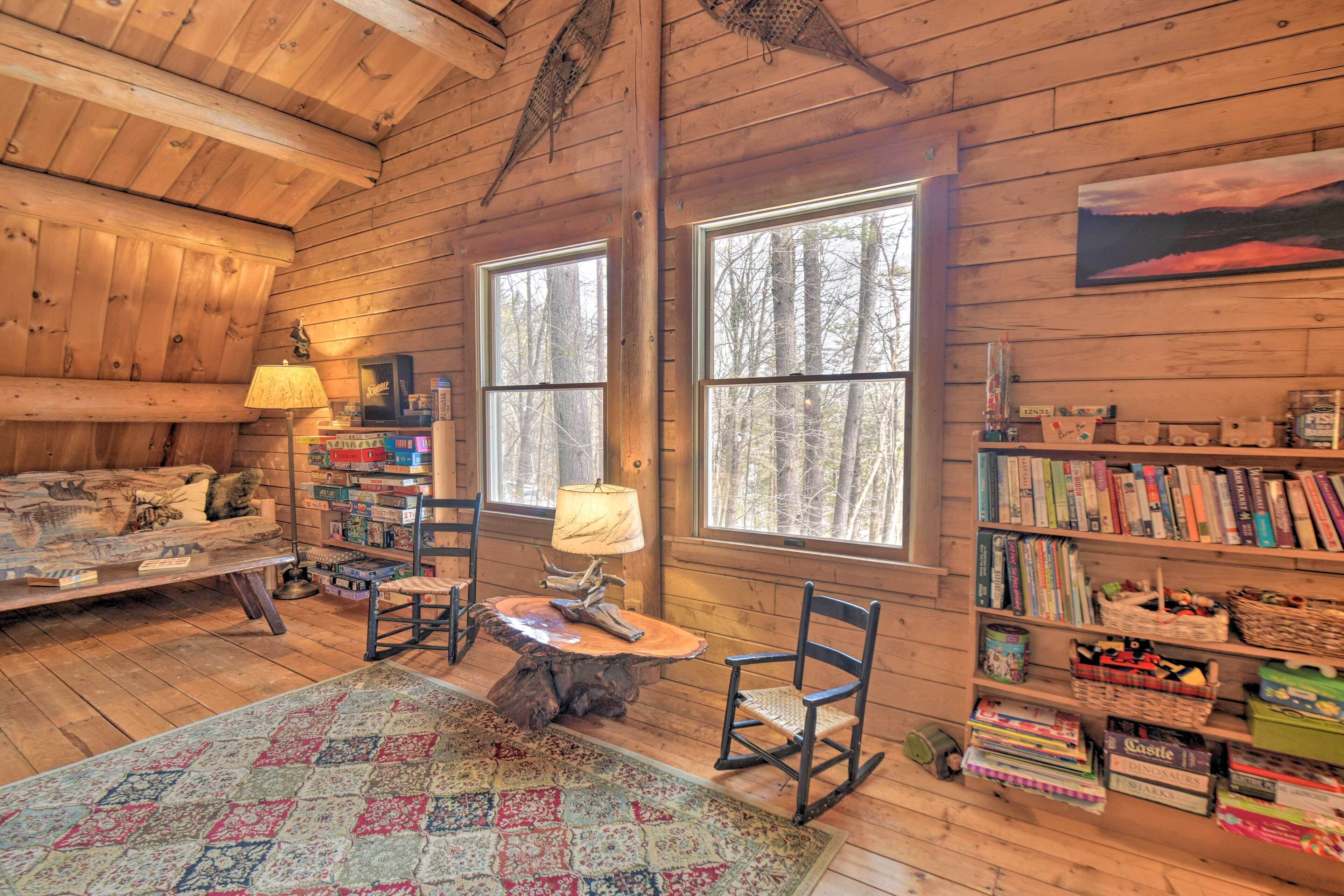 A cozy cabin interior with a bed, a dining table with chairs, a bookshelf, and large windows.