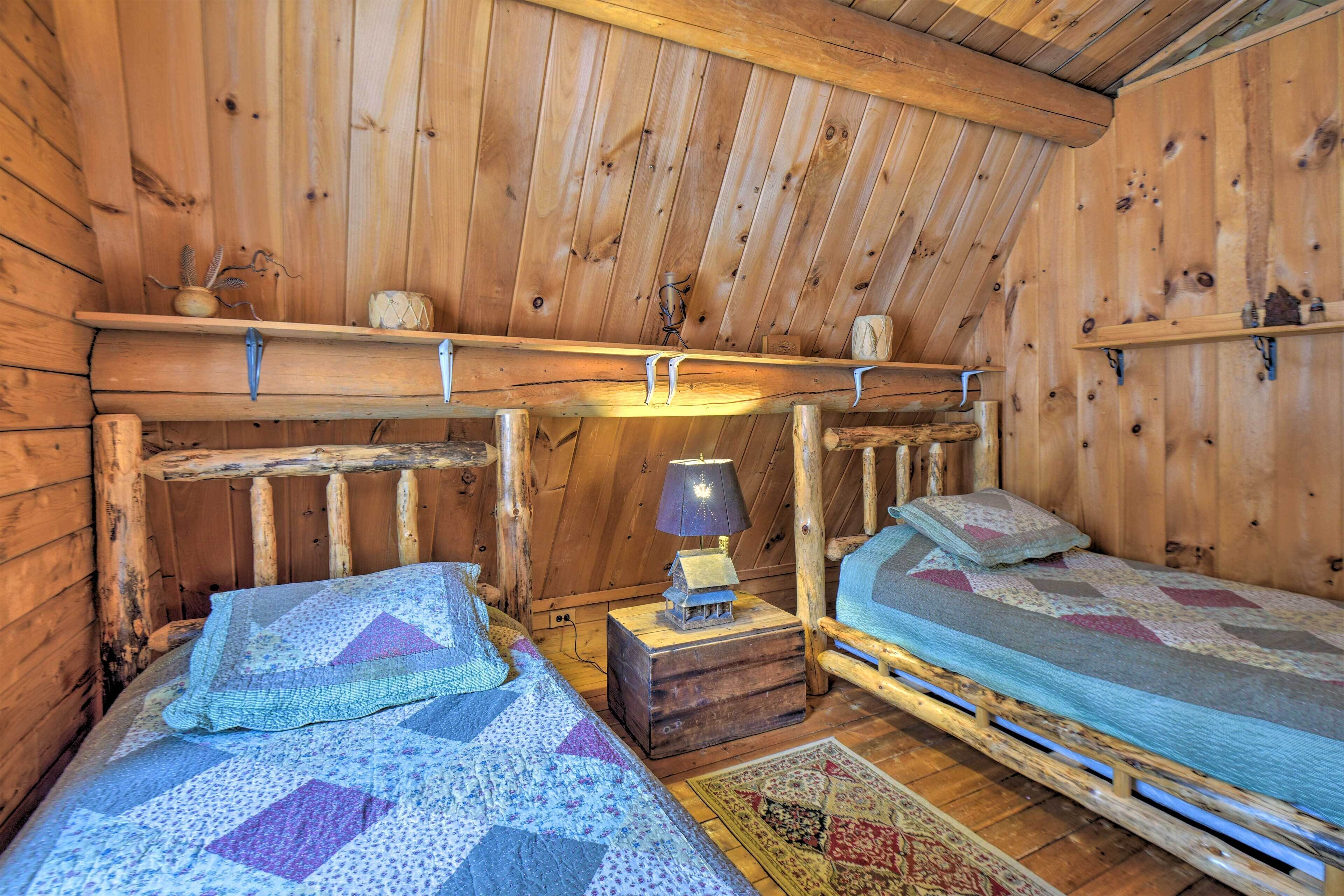 A cozy cabin room with two wooden beds, a small table with a lamp, and a shelf with decorative items.