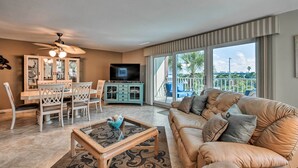A spacious living room with a beige leather sofa, a dining area with white chairs, a television, and a balcony with a view of palm trees.