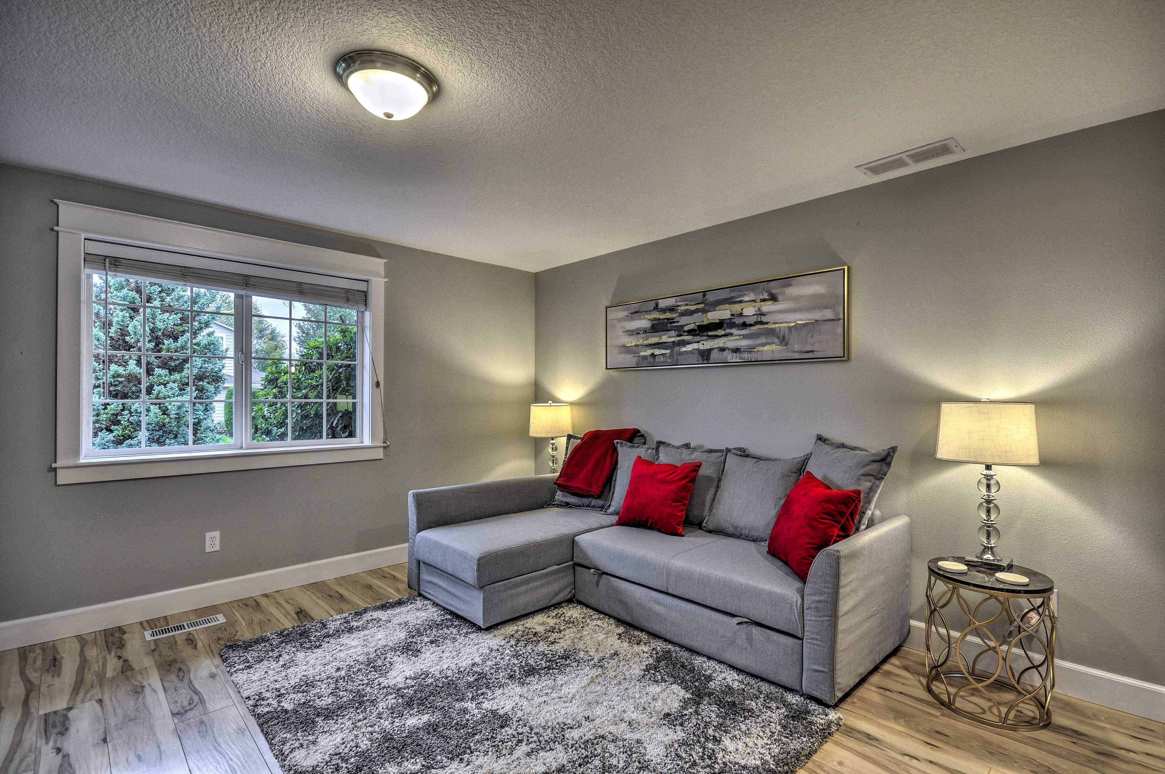 A living room with a grey sectional sofa, red and grey cushions, a black and white rug, a side table with a lamp, and a window with blinds.