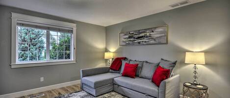 A living room with a grey sectional sofa, red and grey cushions, a black and white rug, a side table with a lamp, and a window with blinds.