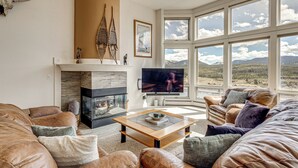 Una sala de estar con sofás de cuero, chimenea, televisor y ventanas amplias con vista a la montaña.