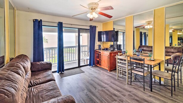 A living room with a brown leather sofa, a wooden dining table with chairs, a kitchen area with a bar, and a large sliding glass door leading to a balcony. - Seaside Corpus Christi Condo at The Villa del Sol! (Corpus Christi)
