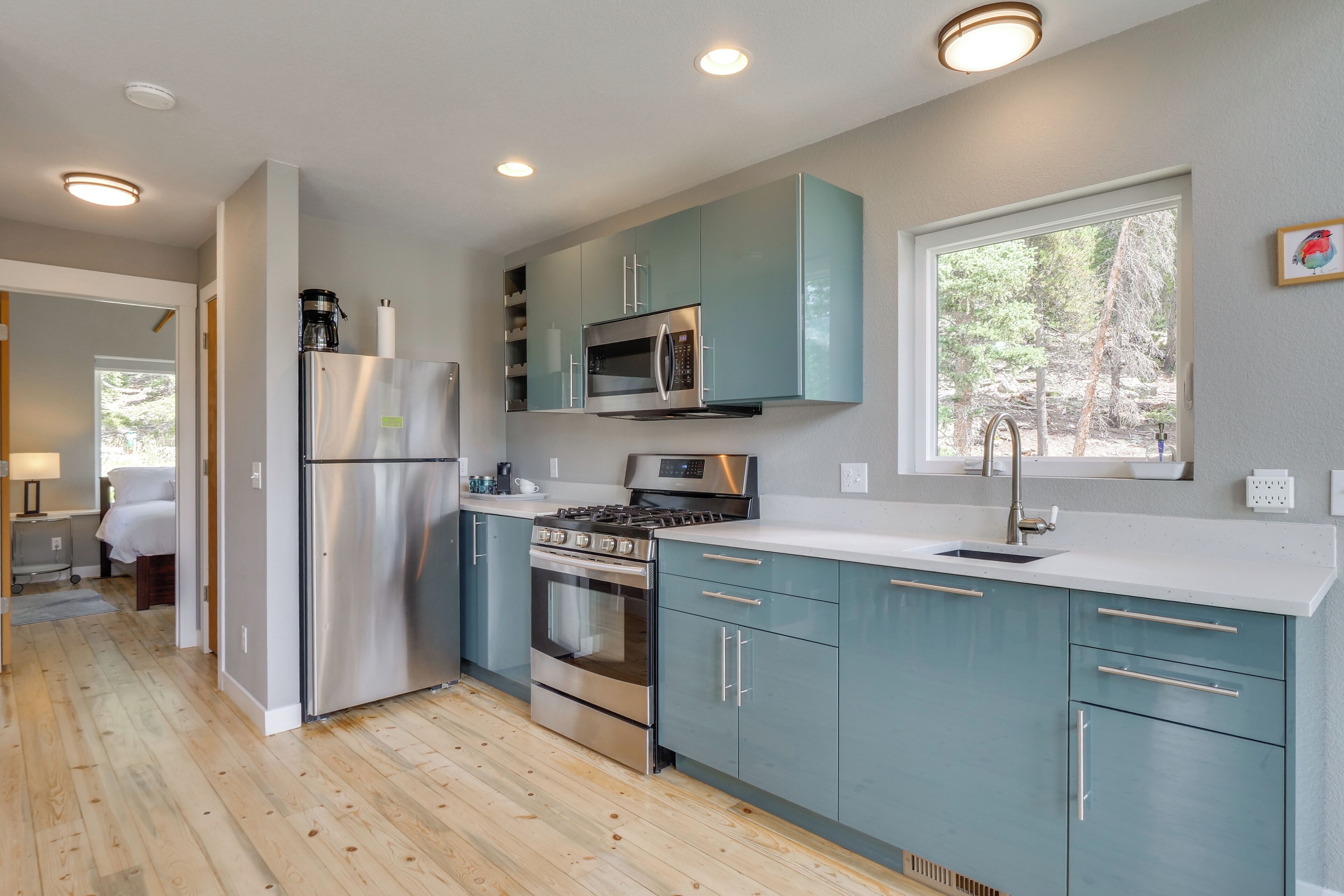 A modern kitchen with stainless steel appliances, teal cabinets, and a large window providing natural light.