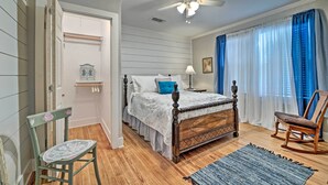 Ein Schlafzimmer mit einem hölzernen Bett, einem Sessel, einem Nachttisch, einem Deckenventilator und einem Fenster mit Vorhängen.