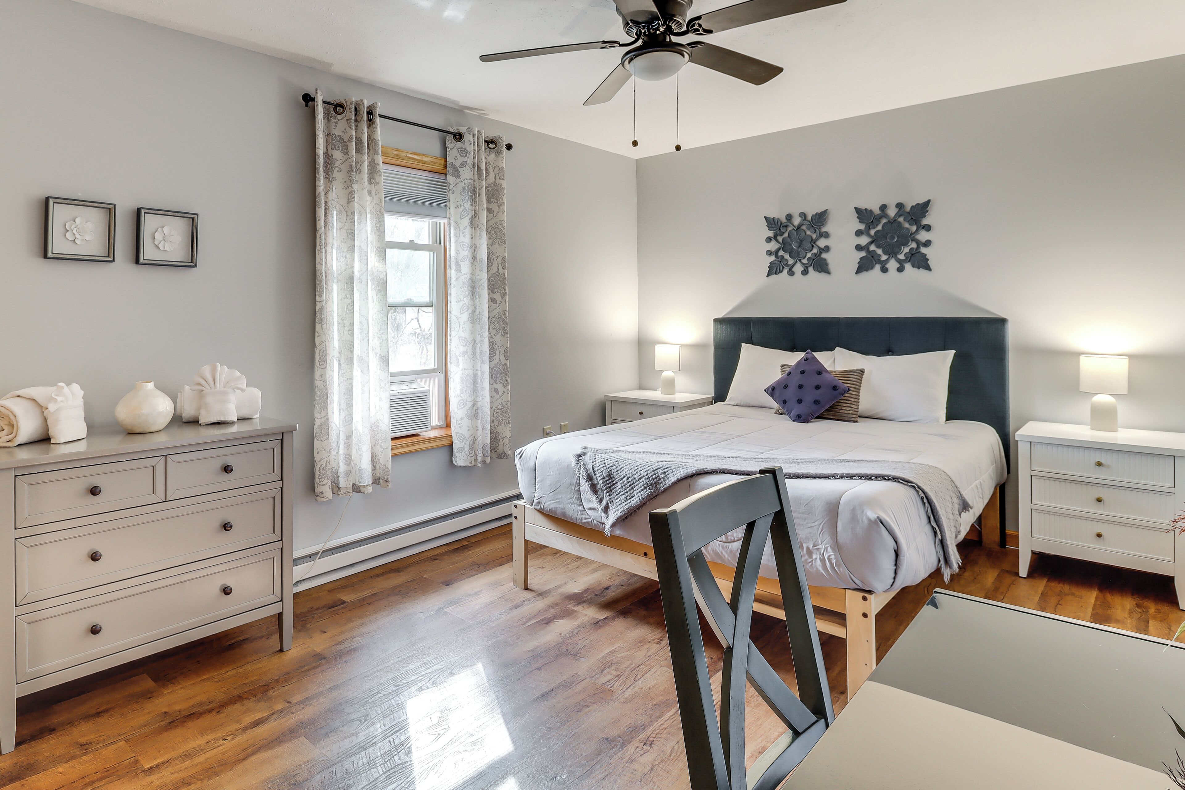 Un dormitorio con cama, mesitas de noche, cómoda, ventilador de techo y ventana con cortinas.