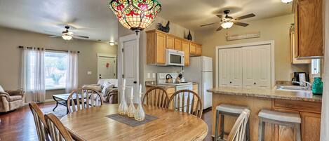 A dining area with a wooden table and chairs, a chandelier, and a kitchen in the background.
