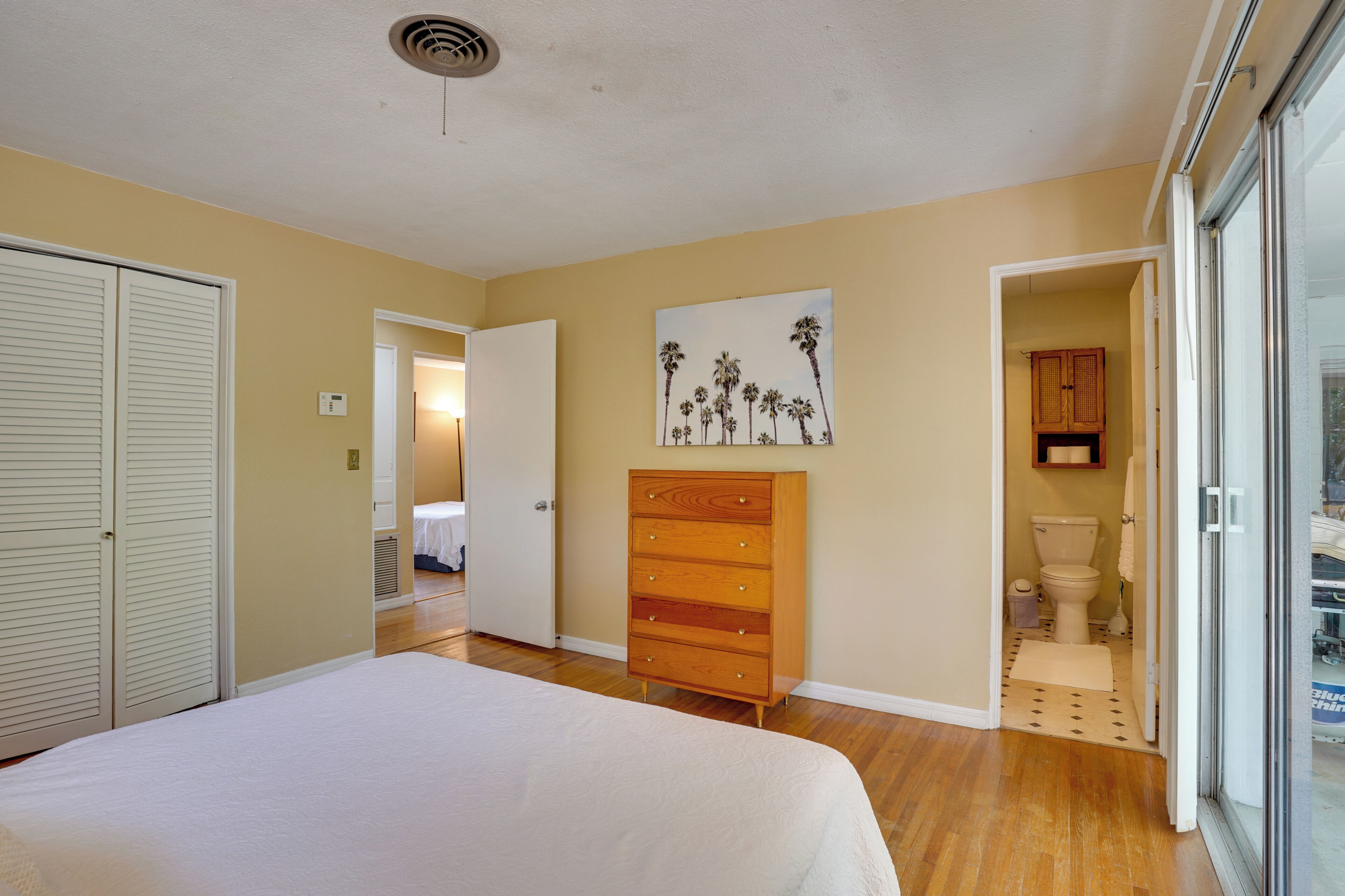 A bedroom with a bed, a wooden dresser, a painting of palm trees, and a bathroom visible through an open door.
