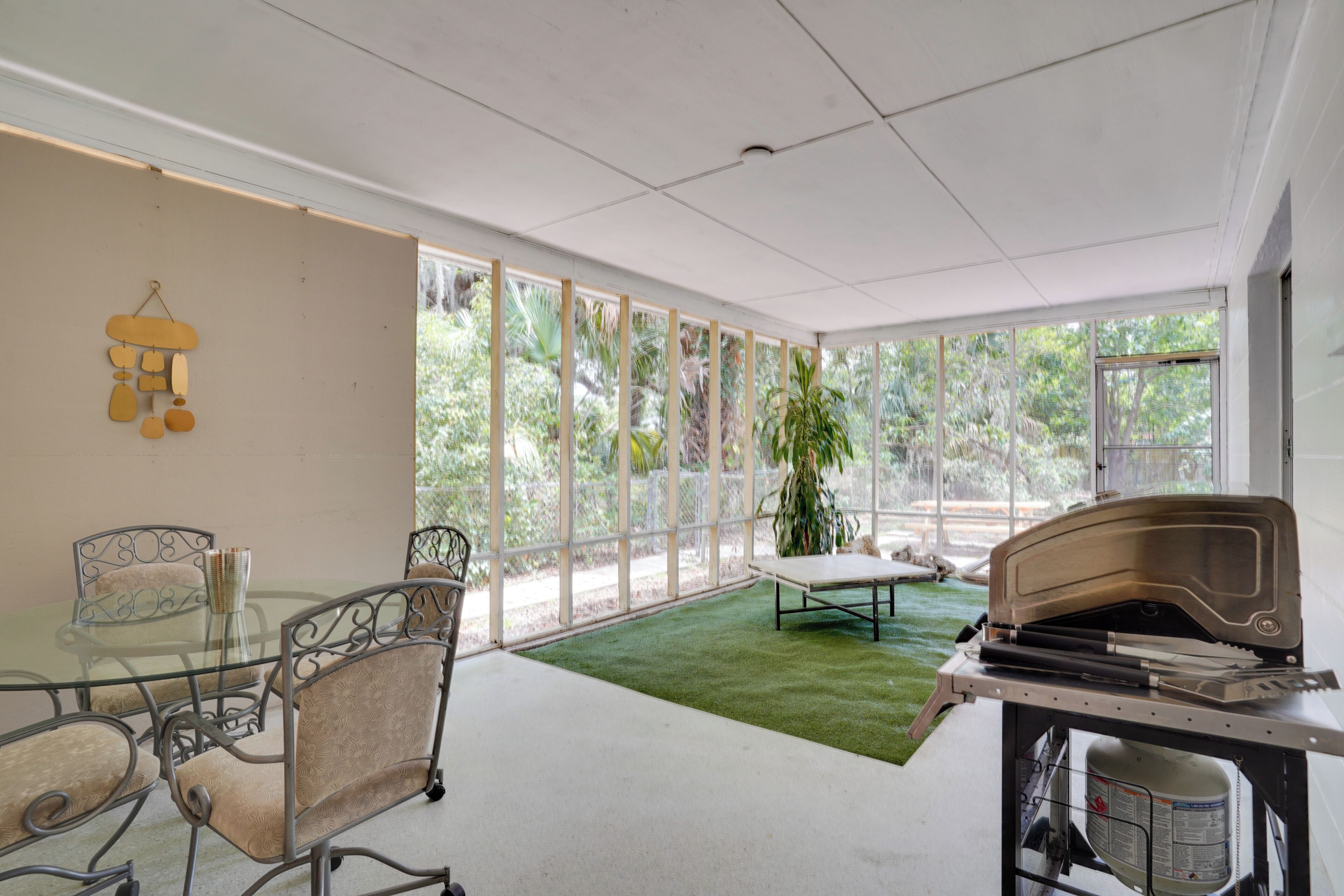 Une pièce spacieuse avec une table en verre, des chaises en métal, un espace recouvert de gazon synthétique vert et de grandes fenêtres offrant une vue sur la verdure environnante.