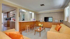 A modern living room with a sofa, coffee table, and bar stools. There is a kitchen area with a refrigerator and a television. - Charming Mid-century House - Just Steps to Lake! (Lakeland)