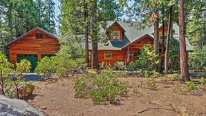 A rustic log cabin with a green garage door, surrounded by dense forest. - Hot Tub, Deck & Trail Access: Shaver Lake Cabin (Shaver Lake)