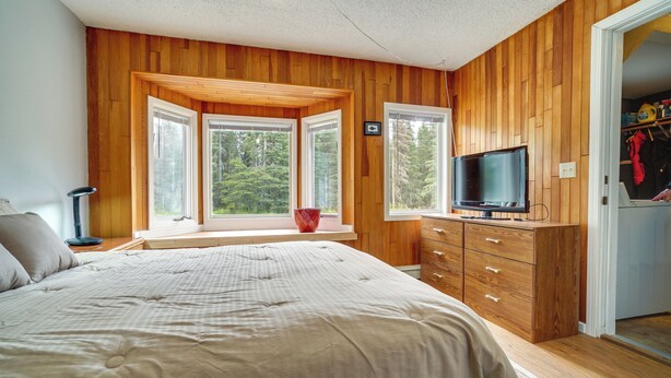Un dormitorio con paneles de madera, una cama grande, un televisor sobre una cómoda de madera y ventanas que dan a un paisaje de árboles.