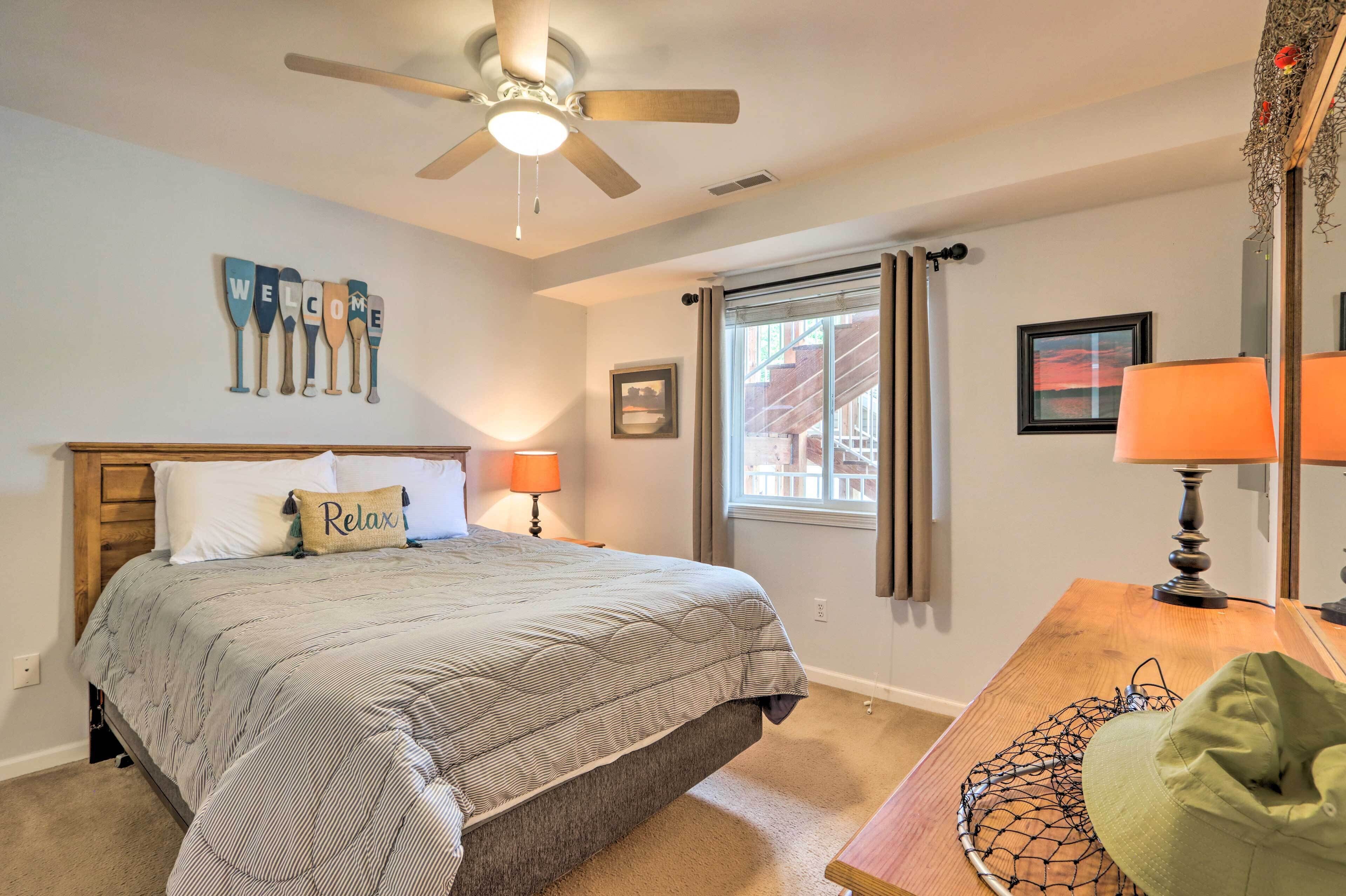A bedroom with a bed, a ceiling fan, a window with curtains, a bedside table with a lamp, and a wall with a 'Welcome' sign.