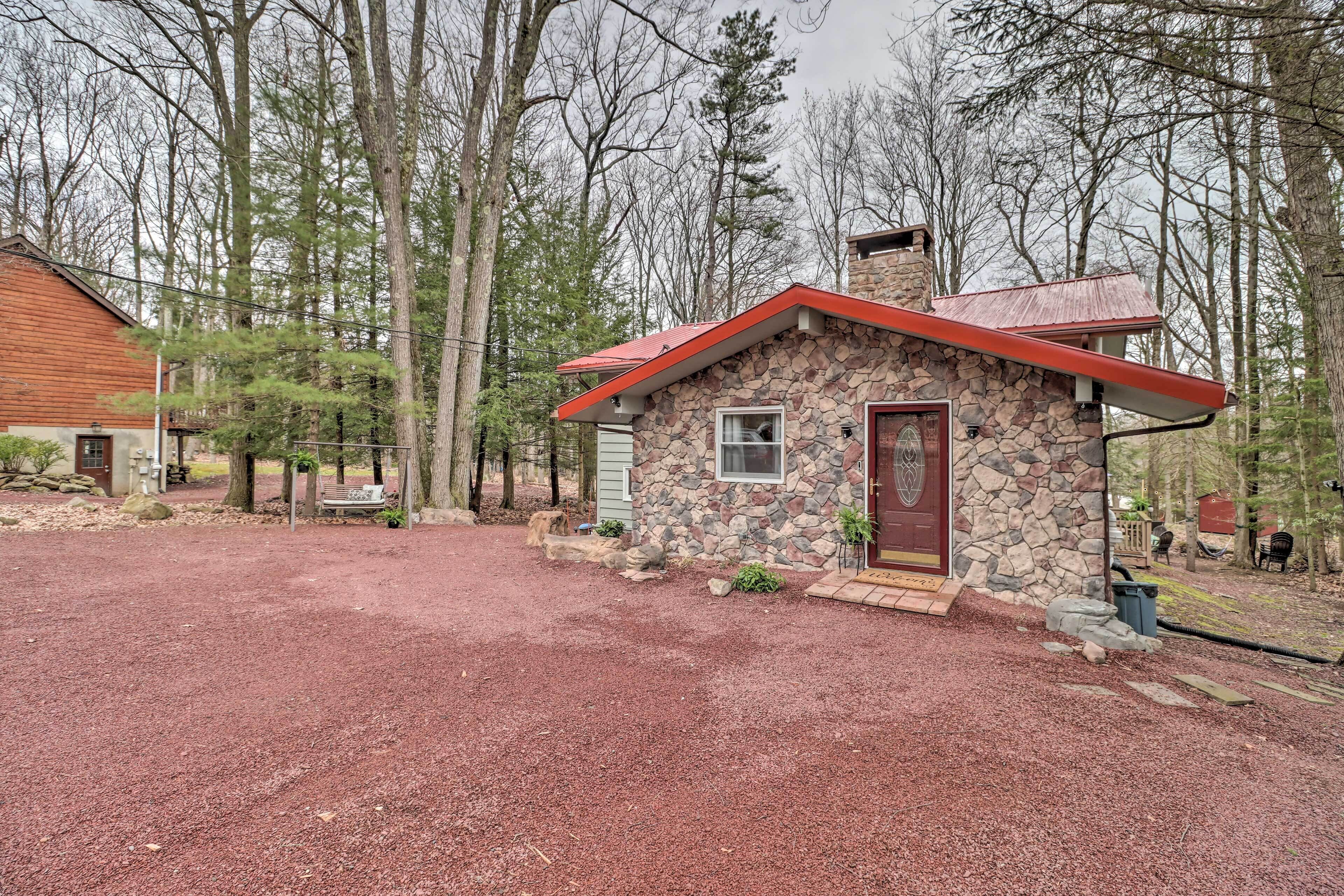 A stone cabin with a red roof, surrounded by a gravel driveway and trees.