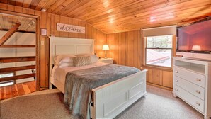 Une chambre confortable avec un plafond en bois, un lit doté d’une tête de lit blanche, une table de chevet avec une lampe, une commode et une fenêtre donnant sur l’extérieur.