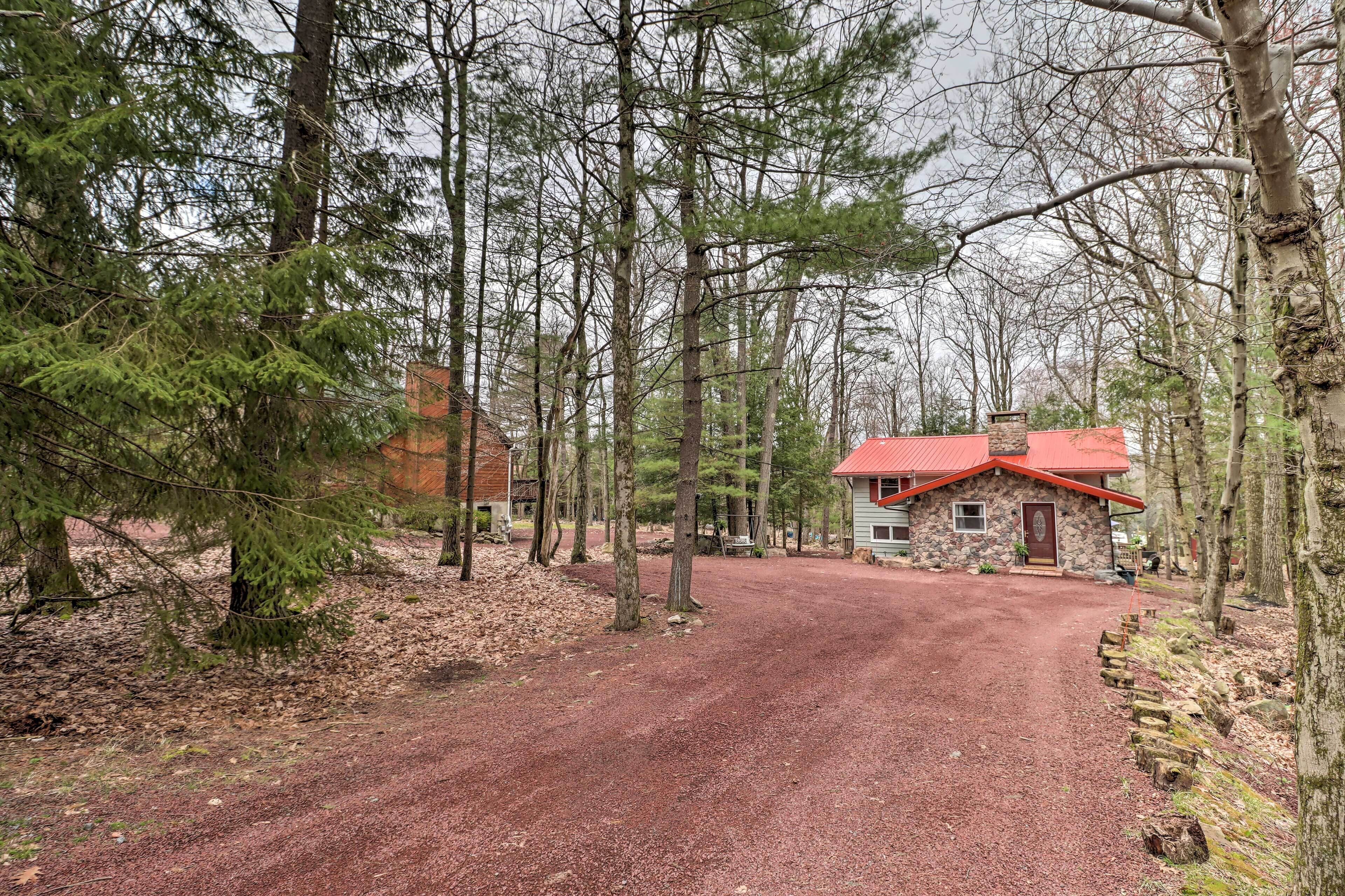 Un chalet rustique avec un toit rouge, entouré de grands arbres et bordé d’une allée en gravier.