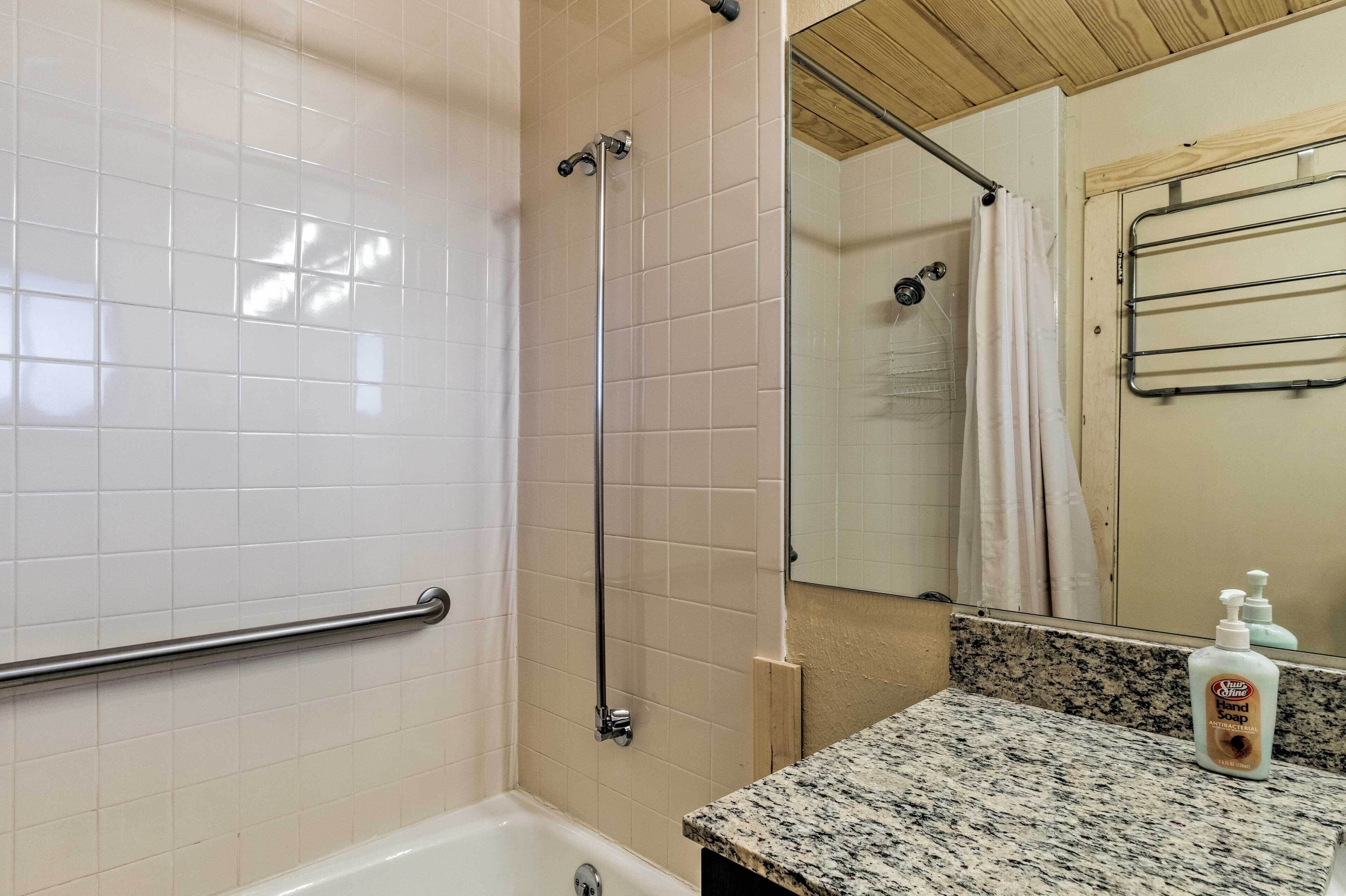 A bathroom with white tiled walls, a granite countertop, a handheld showerhead, a handheld faucet, a towel rack, and a bottle of hand soap.