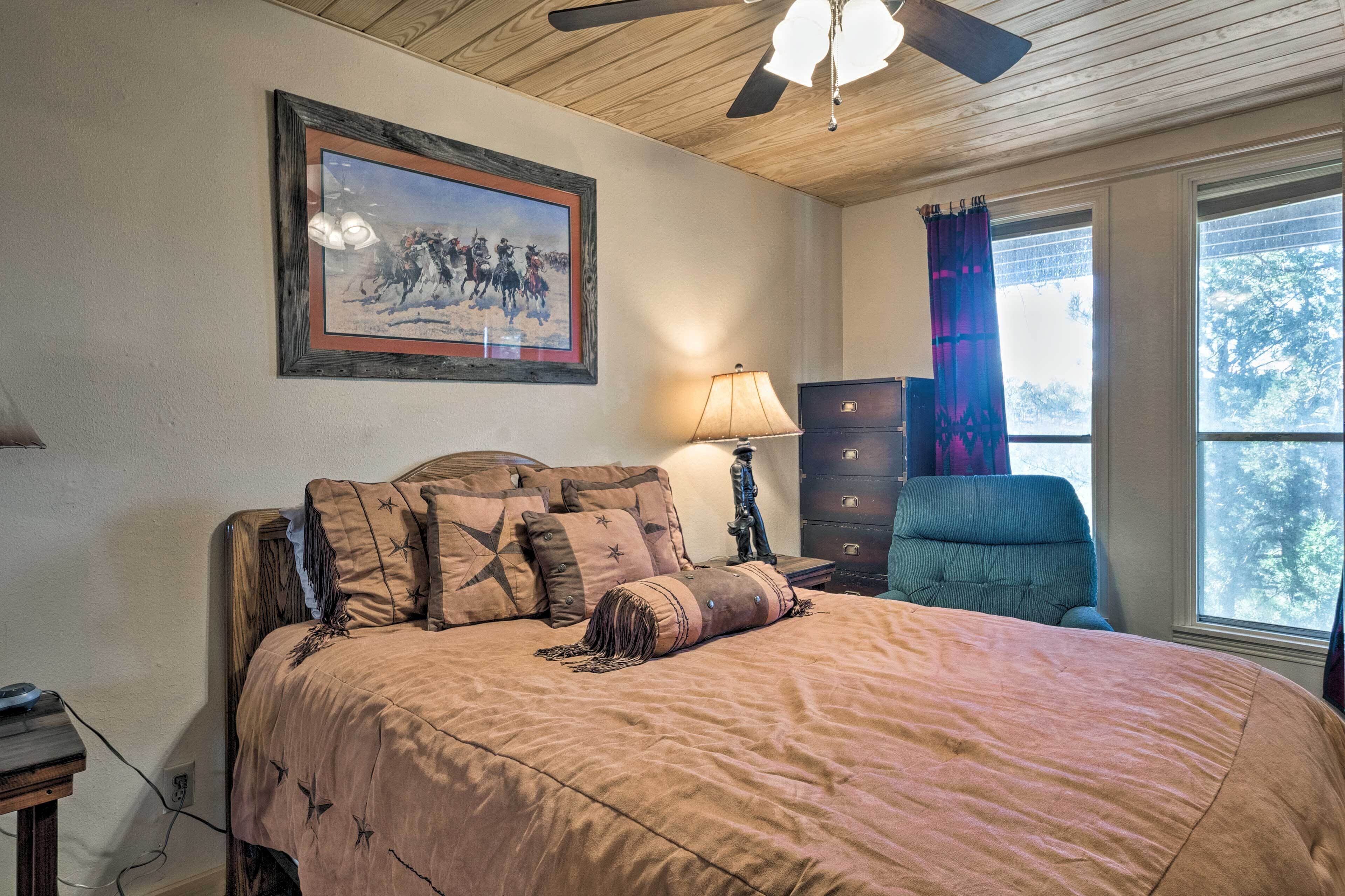 A bedroom with a large bed, a ceiling fan, a framed painting, a dresser, and a window with curtains.