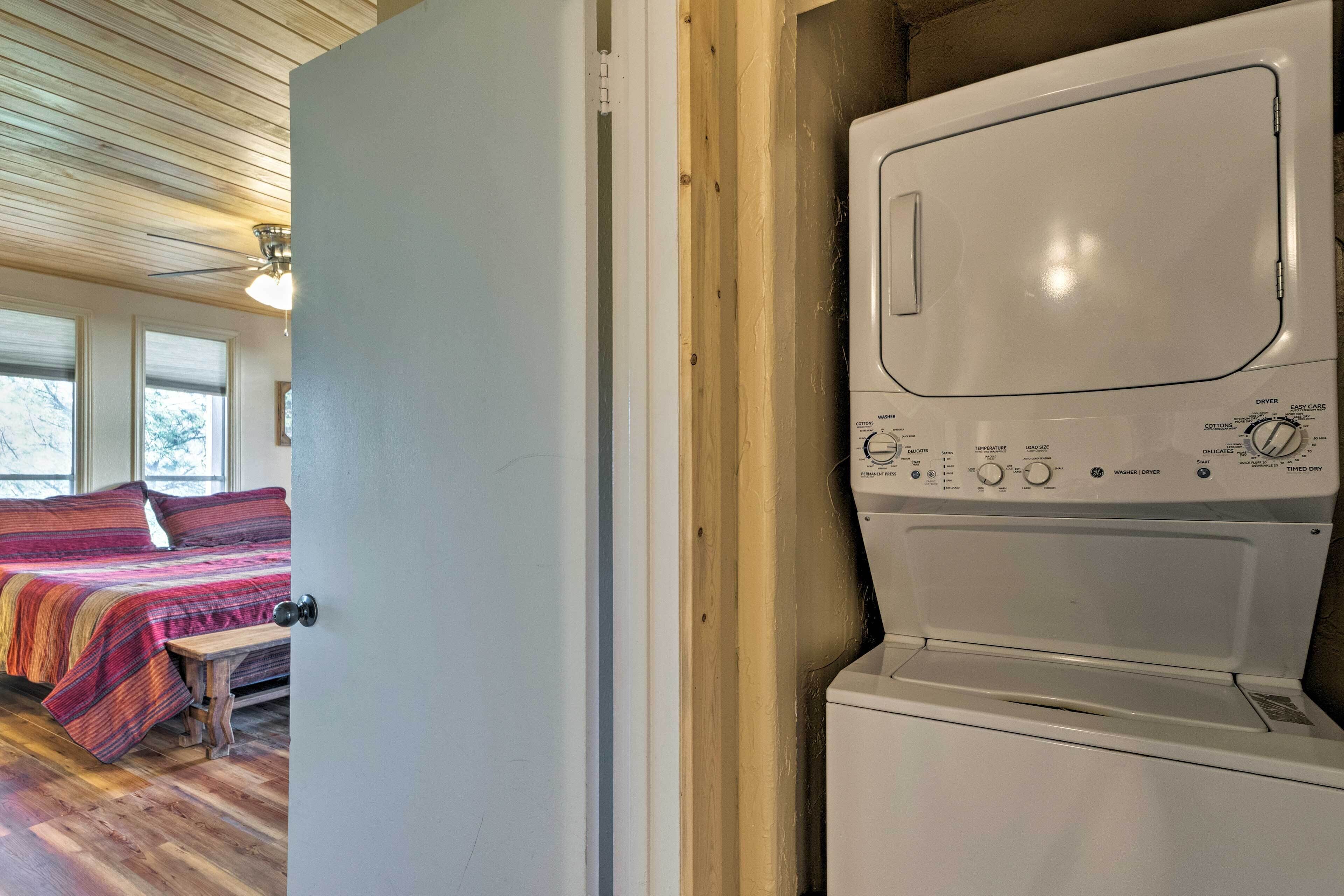 A laundry room with a front-loading washer and dryer, a wooden bench, and a bed with colorful bedding in the adjacent room.