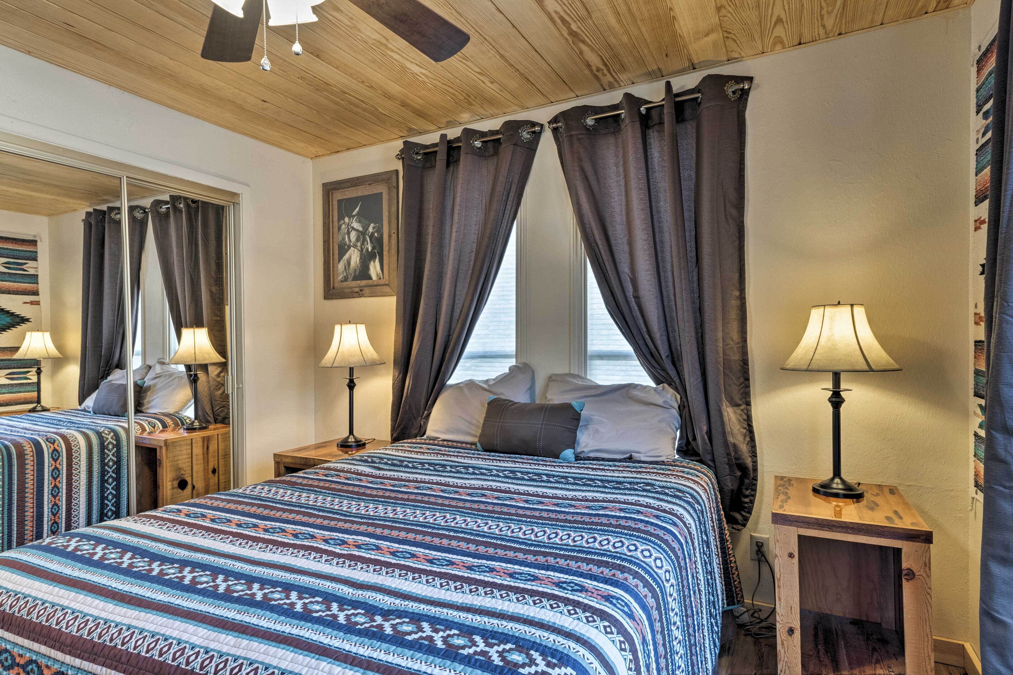 A bedroom with a large bed, two bedside tables with lamps, a ceiling fan, and a view of the sea through curtains.