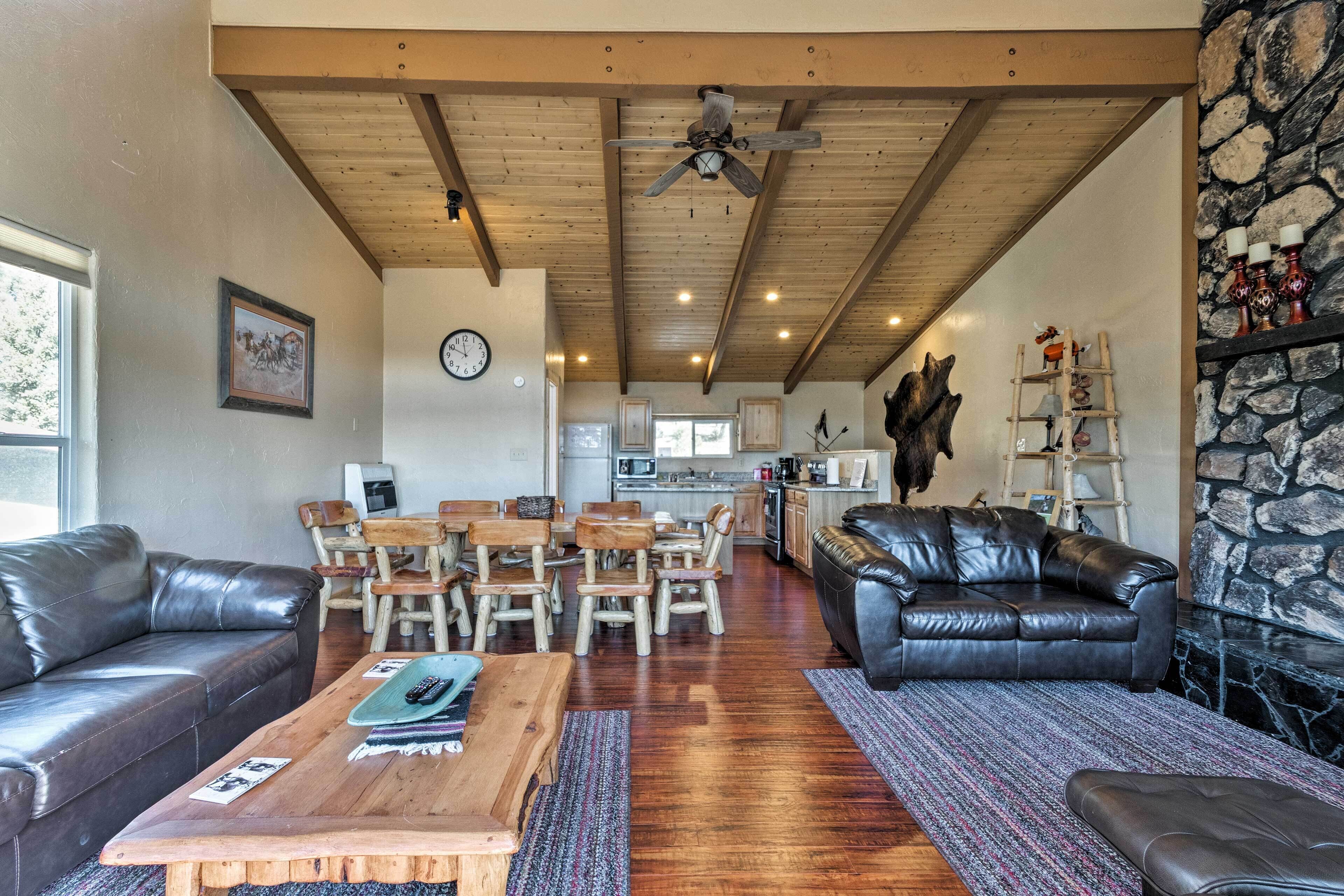 A spacious living room with a wooden ceiling, a stone wall, and a fireplace. There is a leather couch, a coffee table, and a dining area with wooden chairs.