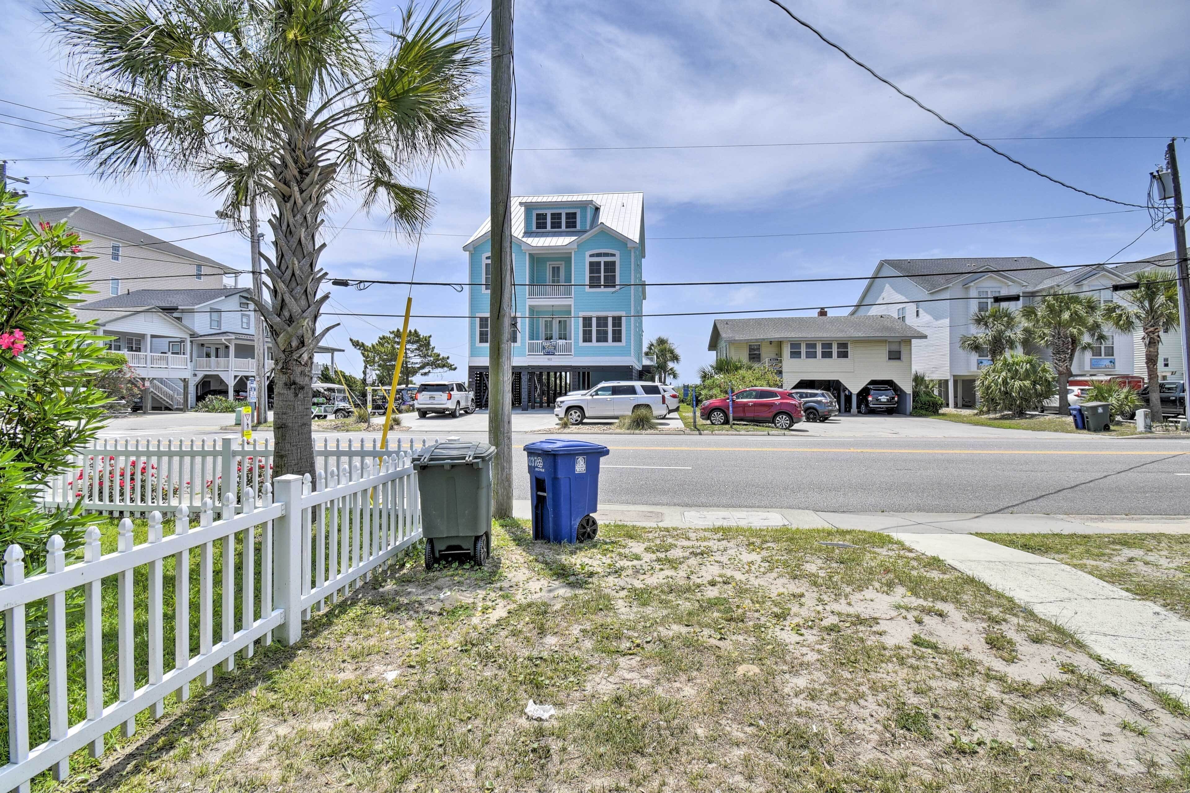 A residential street with houses, a white picket fence, and a blue trash bin.