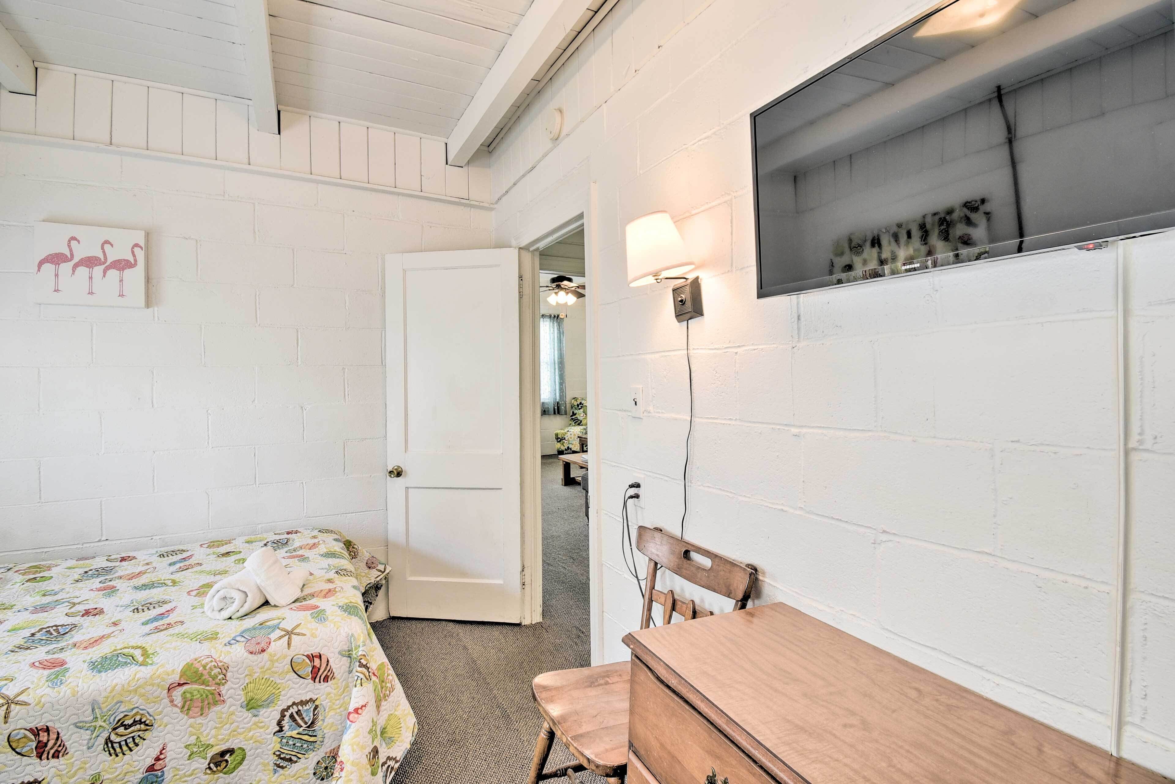 A bedroom with a bed, a wooden dresser, a wall-mounted lamp, and a door leading to another room.