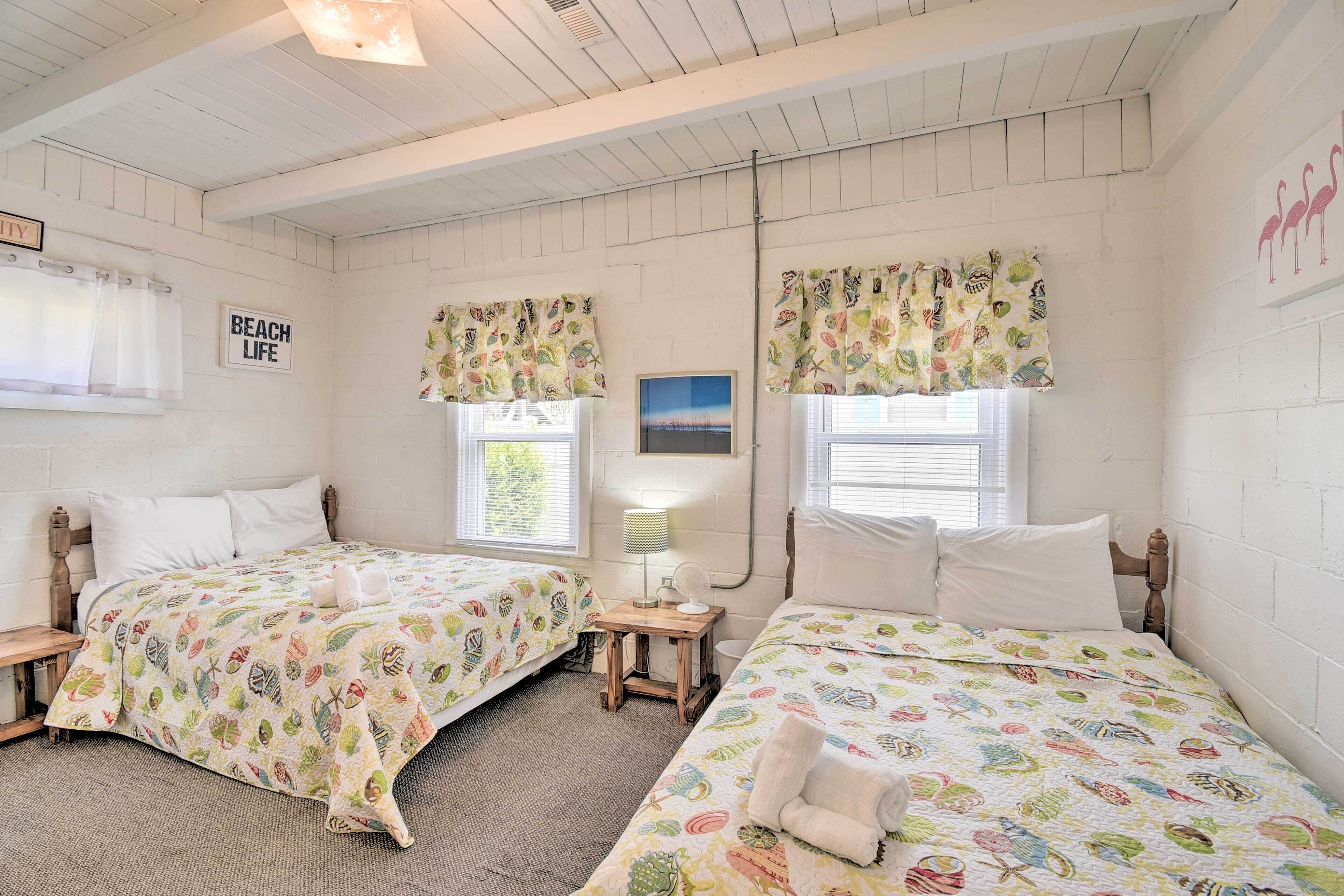 A room with two beds, each with a colorful beach-themed bedspread. There is a small table with a lamp and a picture frame on the wall.