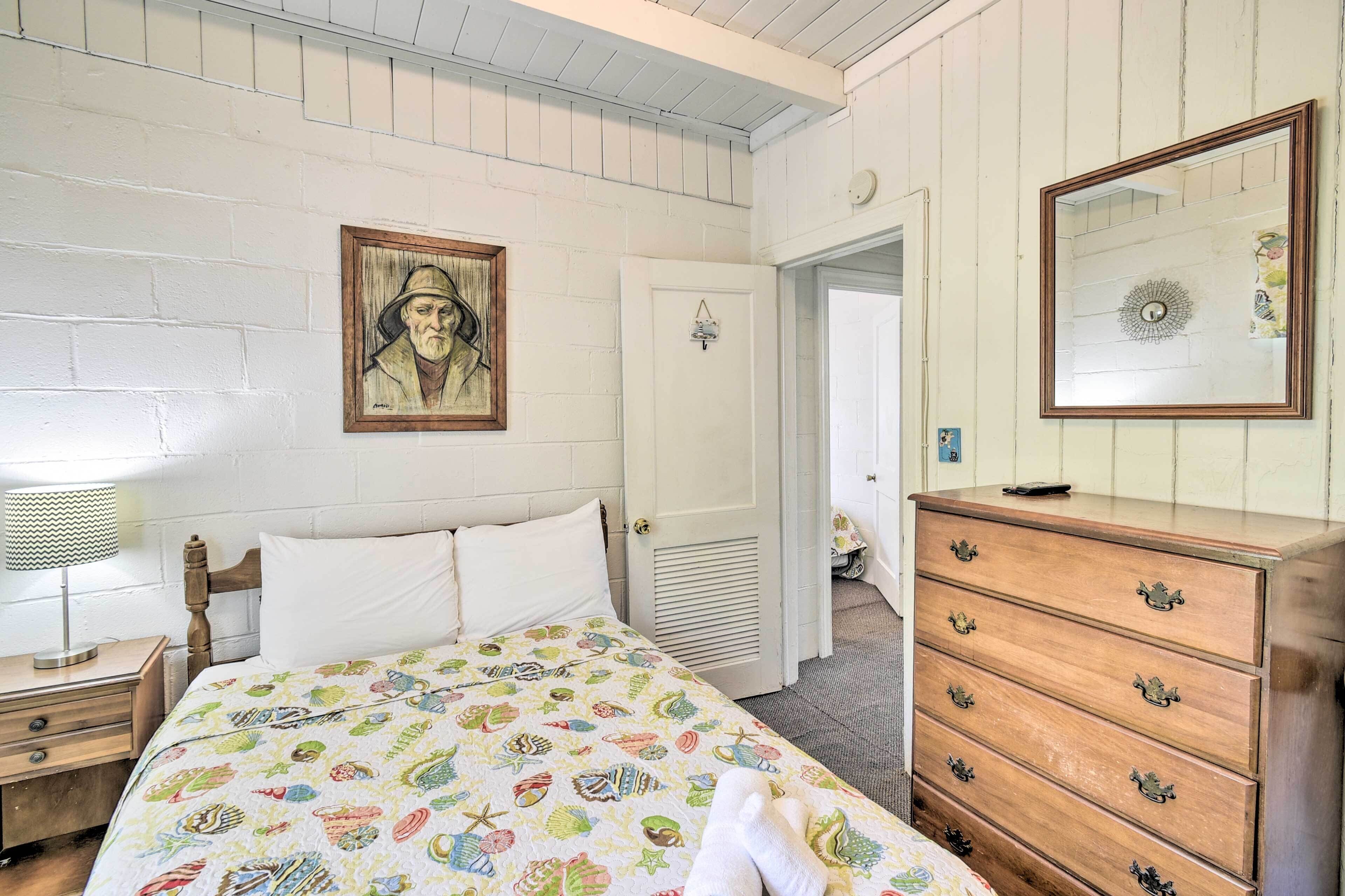Un dormitorio con cama, cómoda, mesita de noche, lámpara, puerta y espejo adosado a la pared.