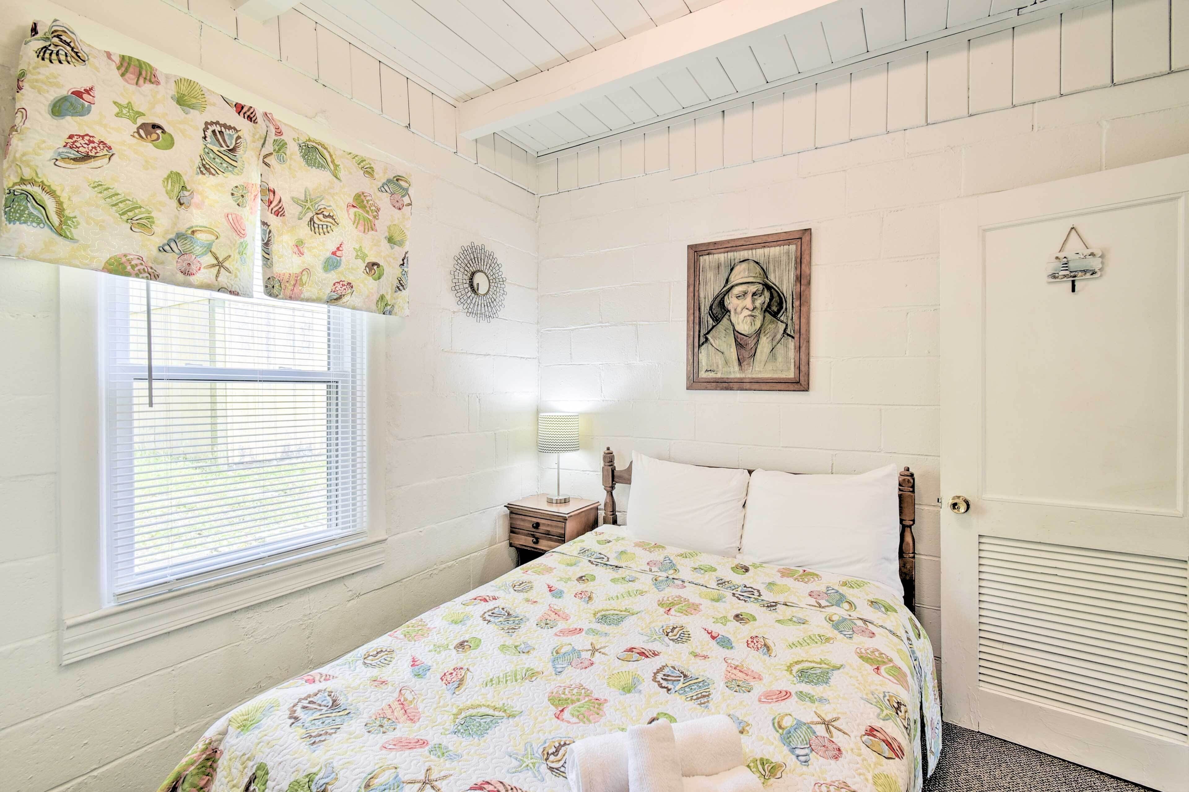 A bedroom with a bed, a window with blinds, a nightstand, a lamp, a wall clock, and a door with a vent.