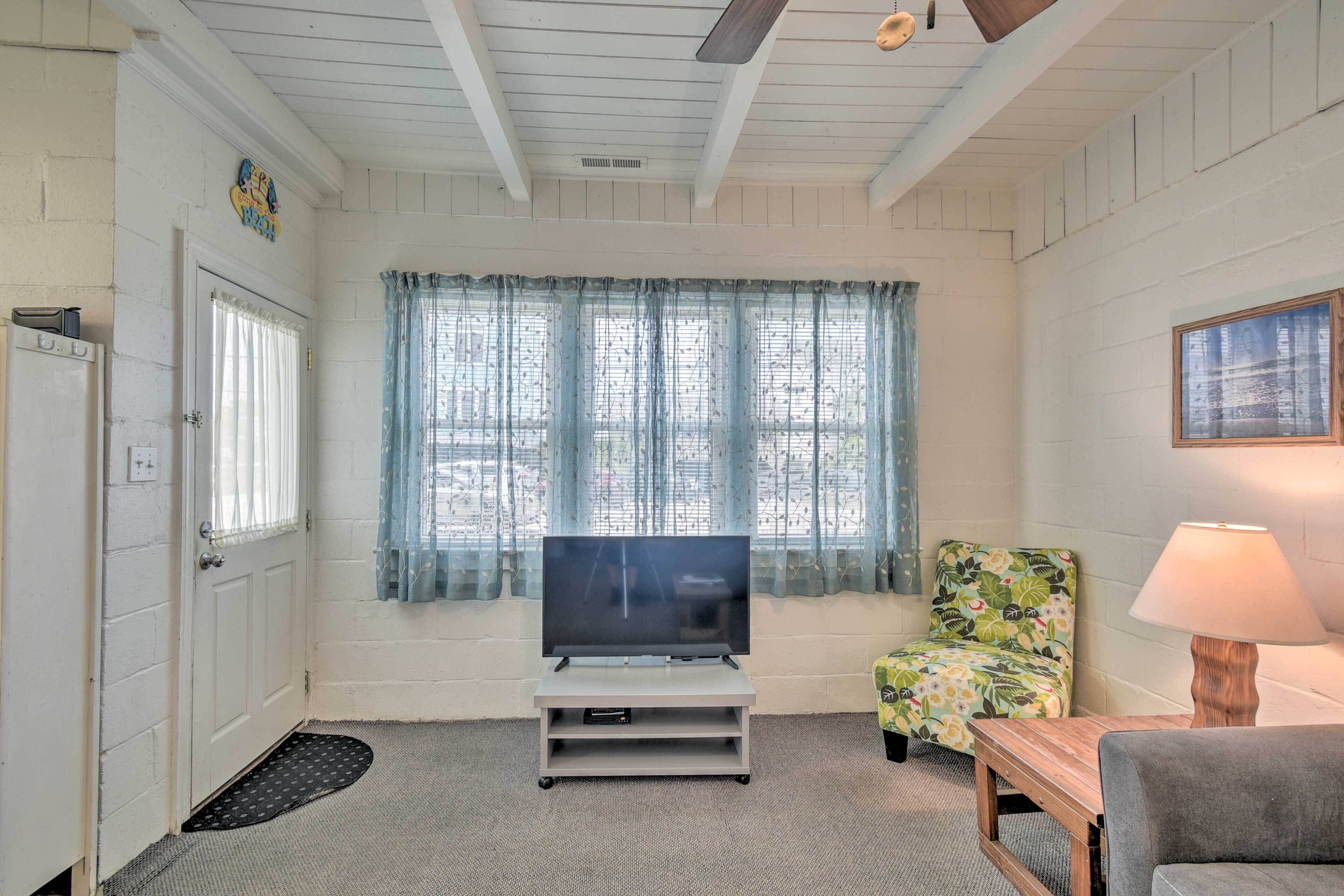 A room with a television on a stand, a floral armchair, a wooden table, and a lamp.