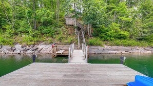 Un muelle de madera que conduce a una pequeña construcción de madera, rodeada de abundante vegetación y un cuerpo de agua tranquilo.