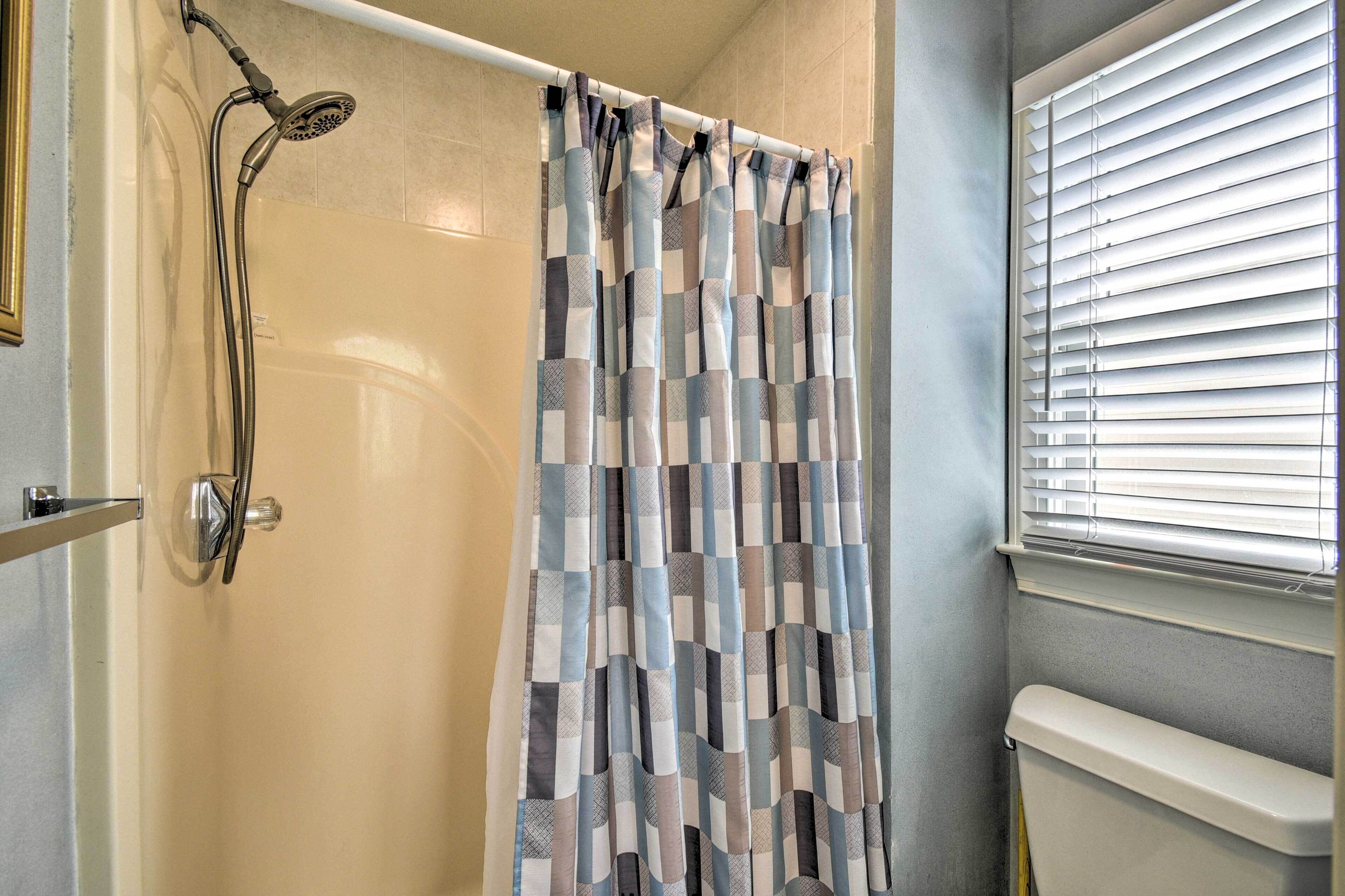 A shower with a checkered curtain, a window with blinds, and a toilet.