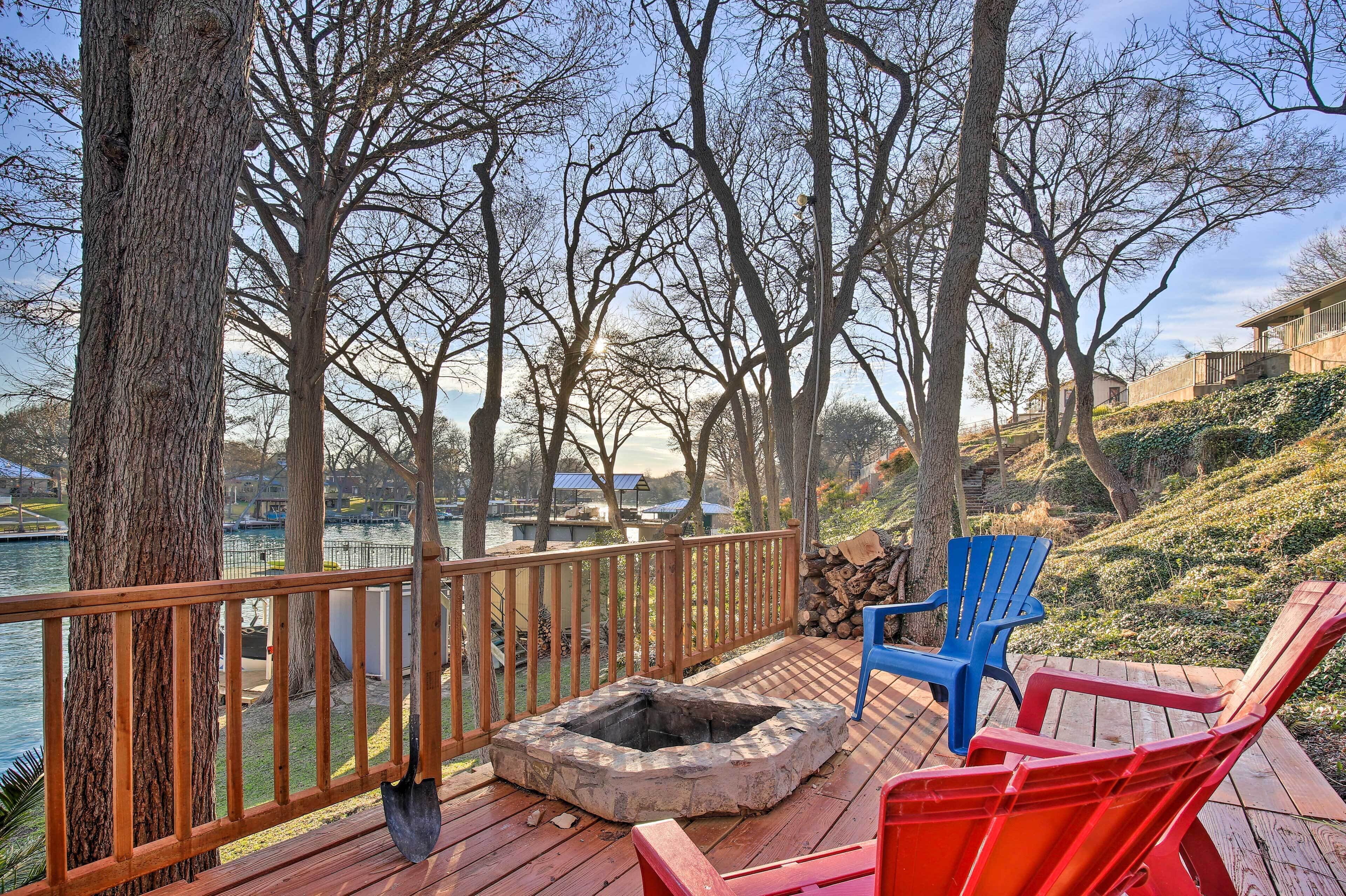 A wooden deck with a stone fire pit, two Adirondack chairs, and a view of a lake with boats.