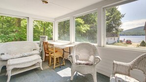 A covered porch with wicker furniture, a table, and chairs, overlooking a lake and forest. - Lake Stinson Cottage w/ Sunroom & Shared Dock! (Rumney)