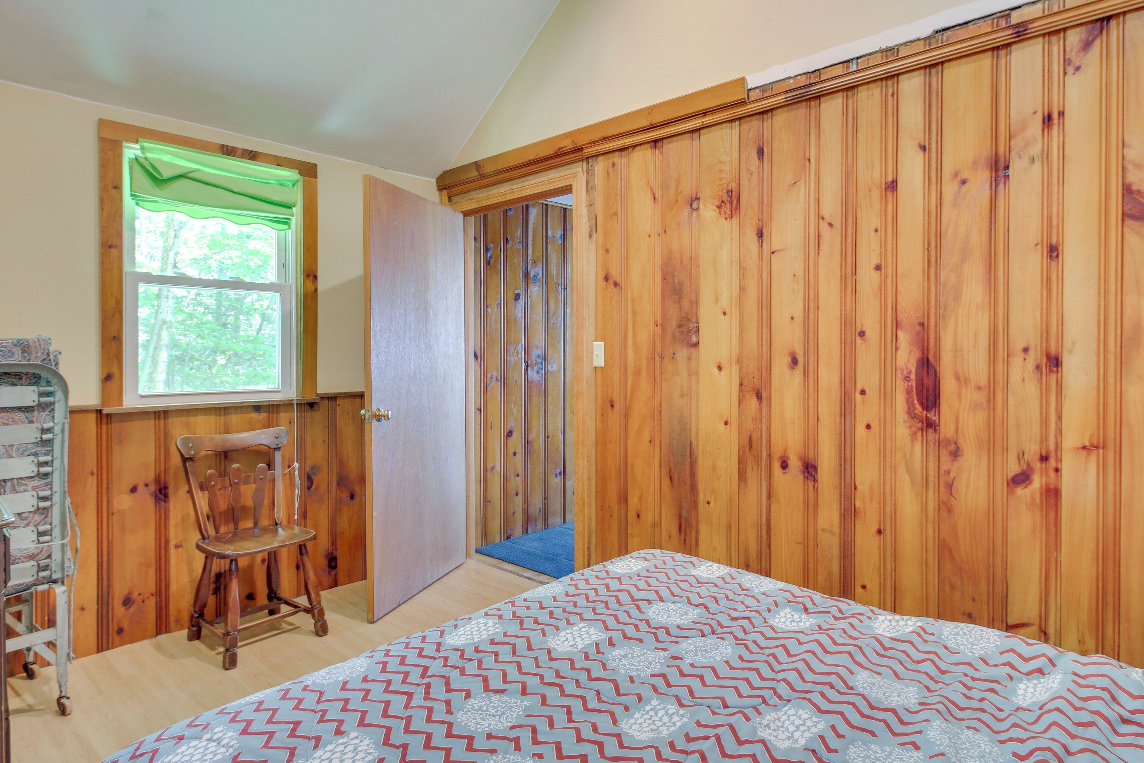 Une chambre avec une armoire en bois, un lit recouvert d’une couvre-lit à motifs, une chaise en bois et une fenêtre agrémentée d’un lambrequin vert.