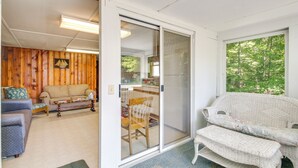 A living room with a sliding glass door leading to a balcony, a wicker sofa, a blue armchair, and a wooden wall with a decorative plate. - Lake Stinson Cottage w/ Sunroom & Shared Dock! (Rumney)