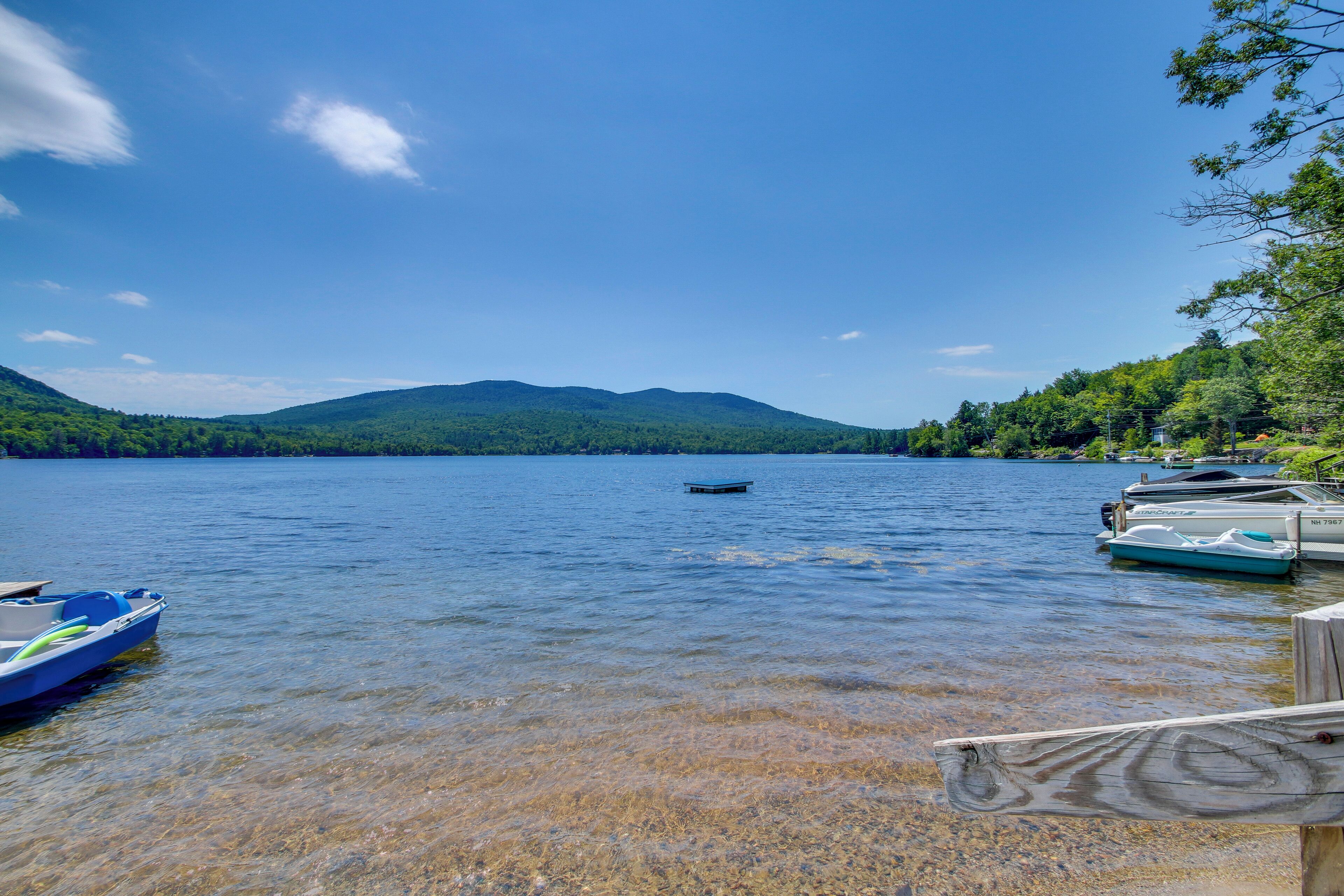 A calm lake with boats docked on the shore, surrounded by forested hills.