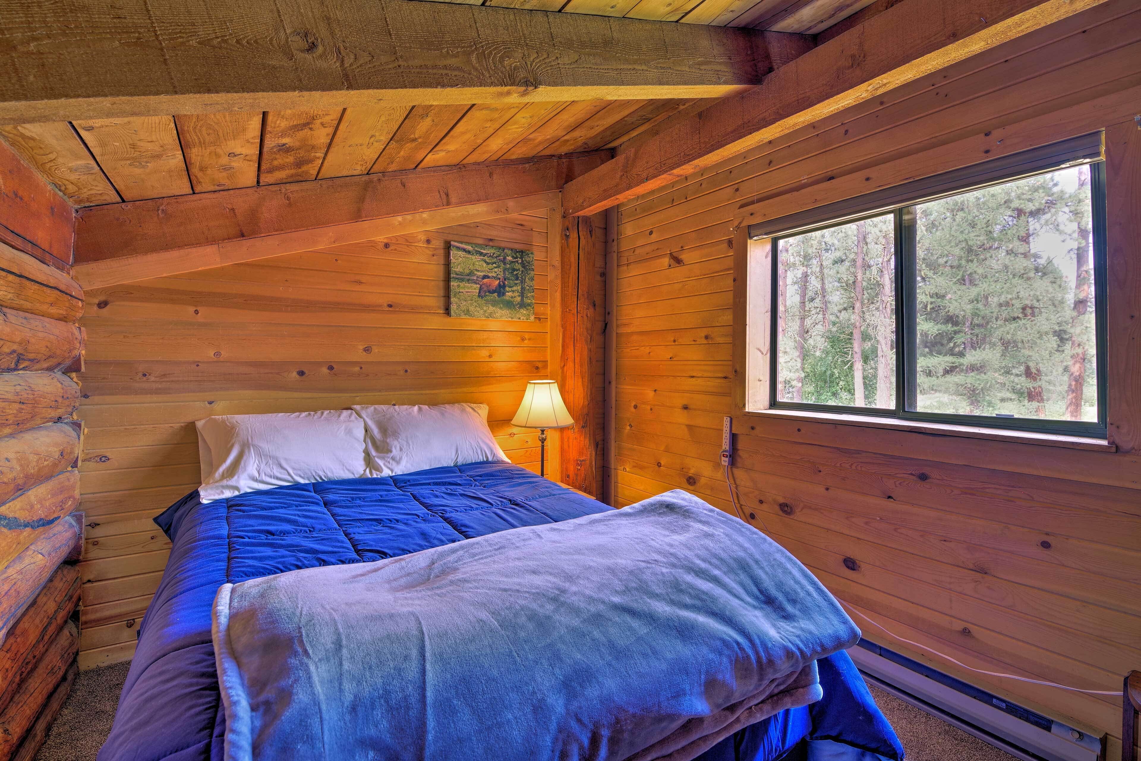 A log cabin bedroom with a bed, a nightstand, a lamp, and a window with a forest view.