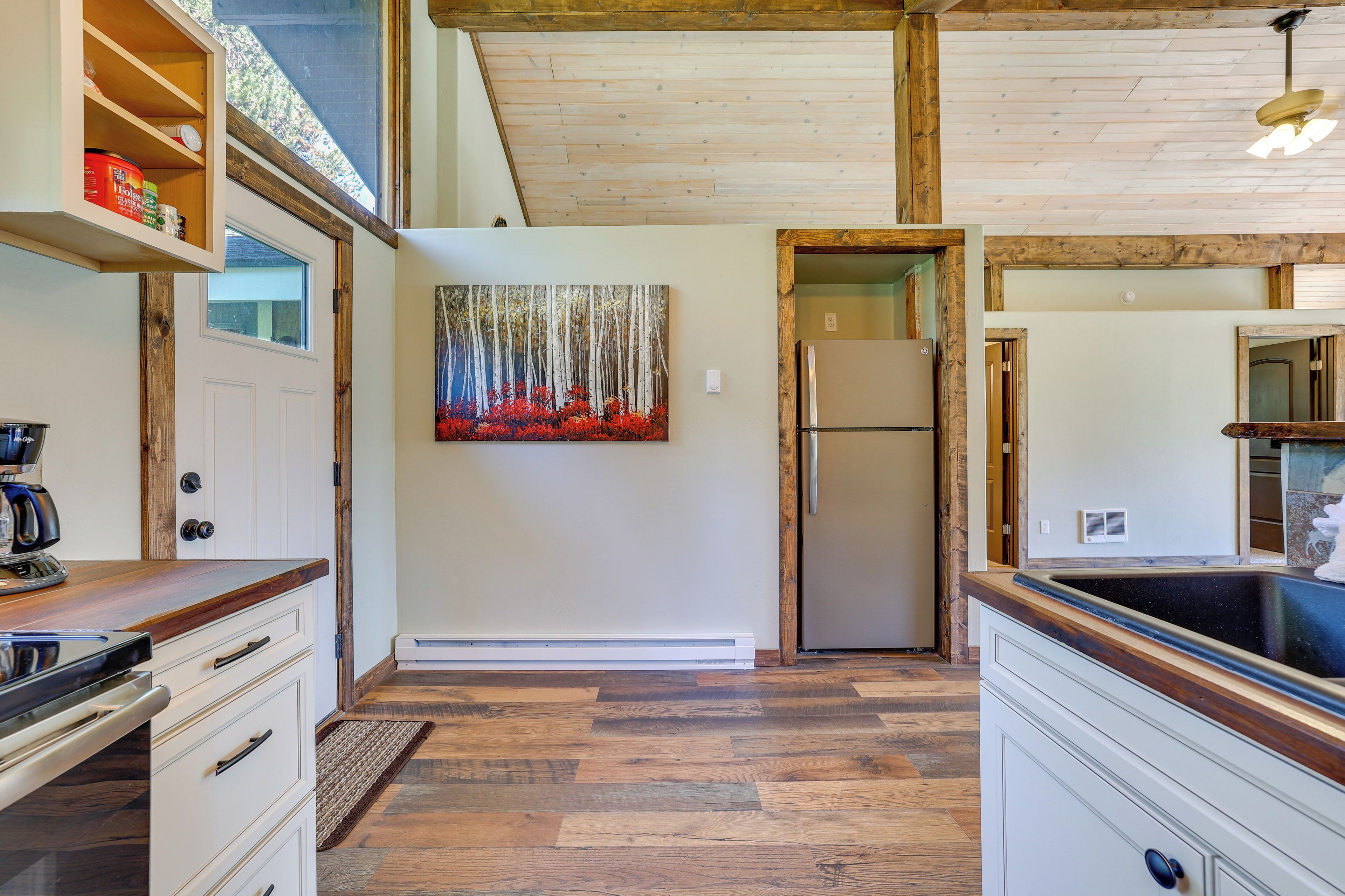 A kitchen with wooden flooring, white cabinetry, a refrigerator, and a painting of trees and flowers on the wall.