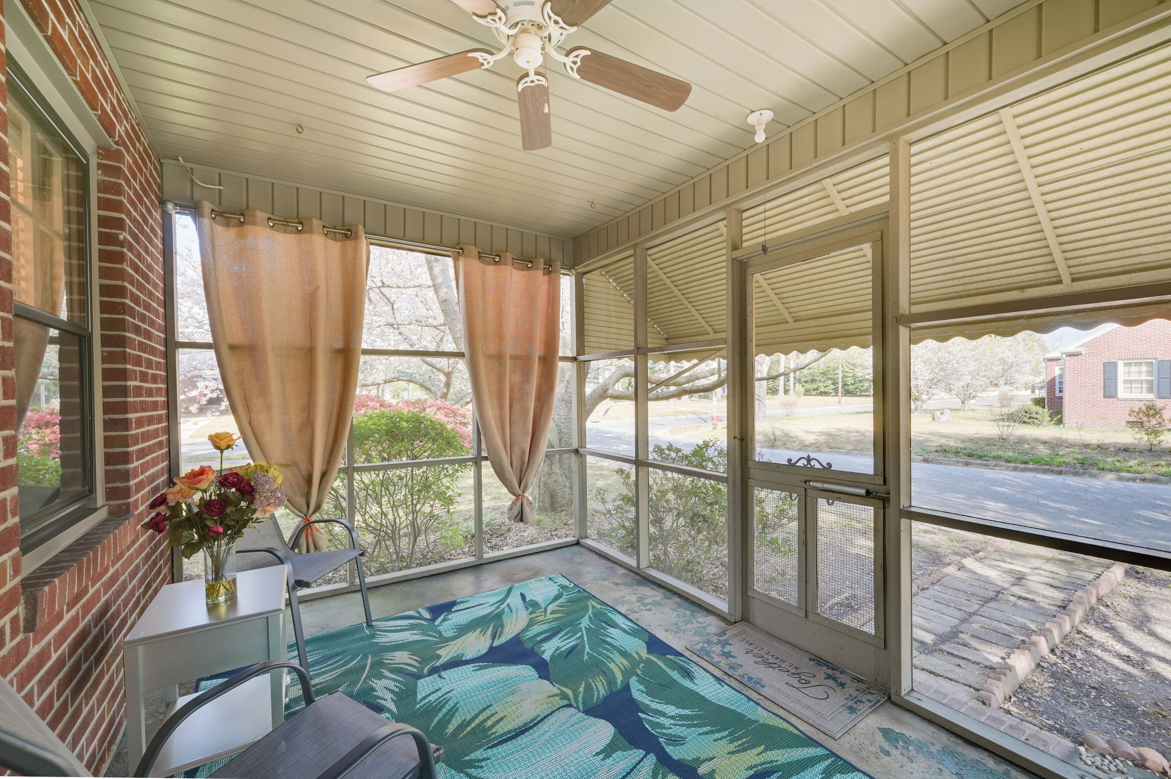 A covered porch with a ceiling fan, a table with chairs, a vase of flowers, and a view of the street.