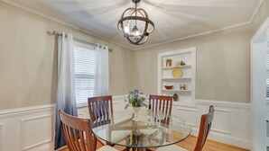 A dining room with a glass table, wooden chairs, a chandelier, a window with curtains, and a shelf with decorative items. - Quaint Goldsboro Home w/ Screened-in Porch! (Goldsboro)