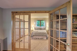 A bedroom with a large wooden door leading to a bathroom, a bed with a grey bedspread, and a wooden bookshelf with books and decorative items.
