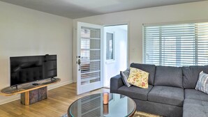 A living room with a flat-screen TV on a wooden stand, a glass coffee table, a grey sofa with floral cushions, and a door with a glass panel. - Florida Home ~ 10 Mi to St Pete Beach! (St. Petersburg)