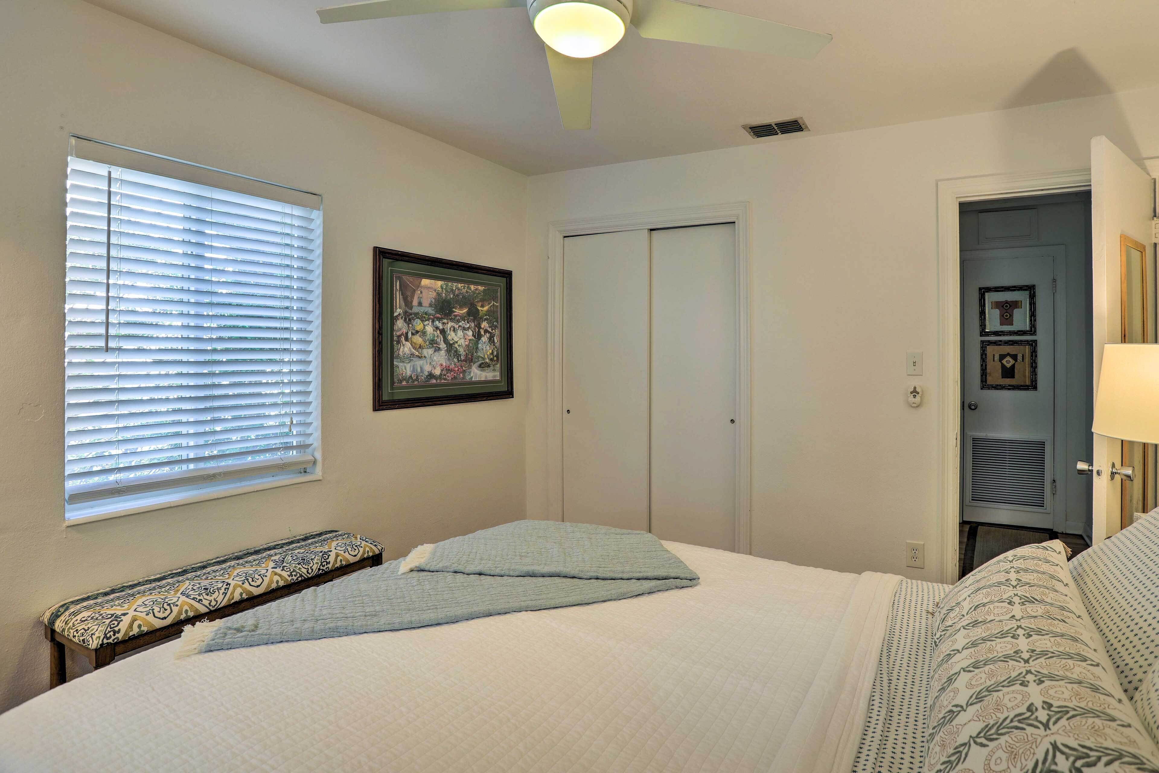 A bedroom with a bed, a bench, a ceiling fan, a window with blinds, and a door.